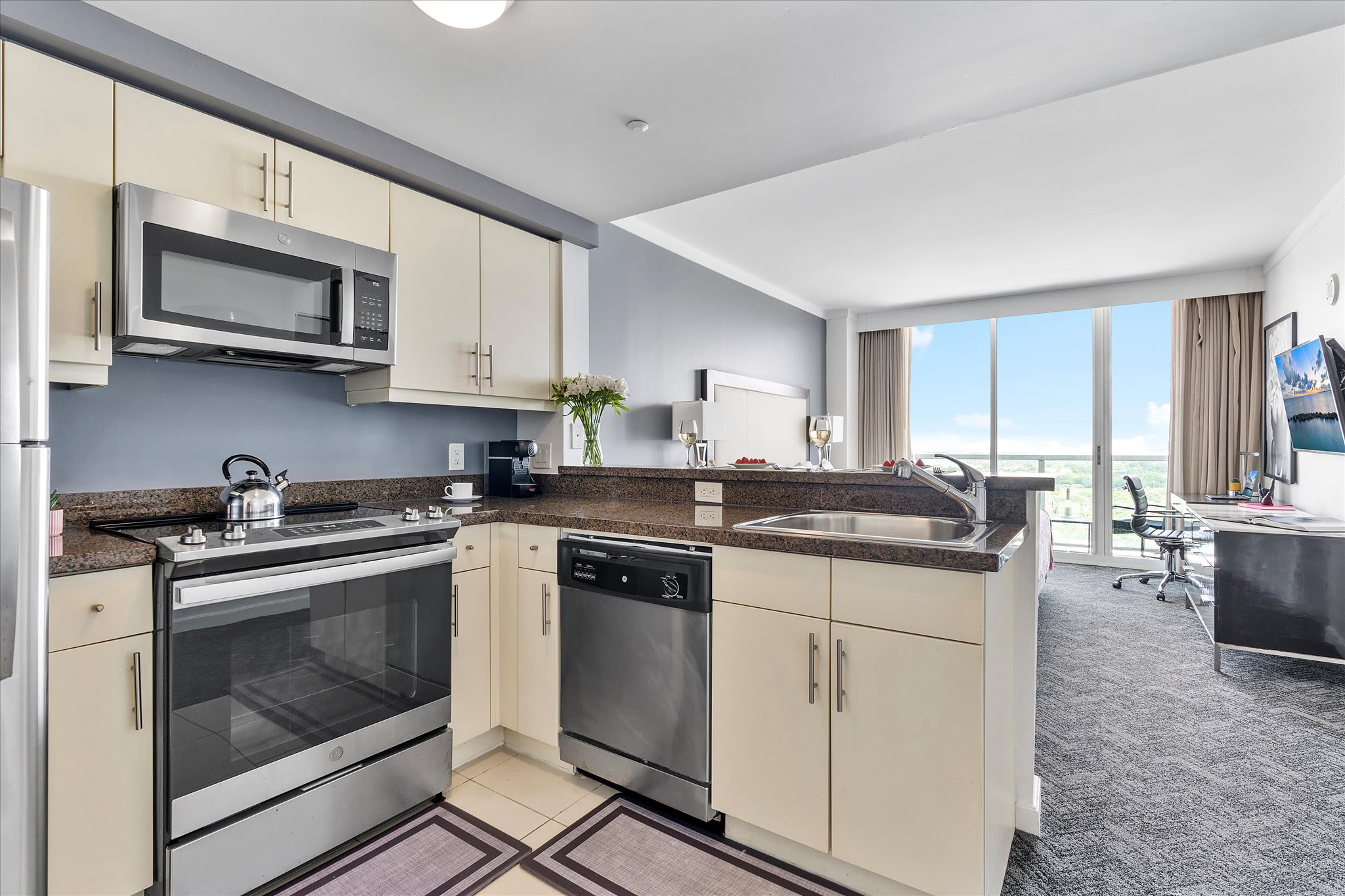Compact modern kitchen with light beige cabinets, stainless steel appliances including a refrigerator, microwave, oven, and dishwasher. Dark granite countertops and a stainless steel sink are visible, with a view into the adjacent living area.