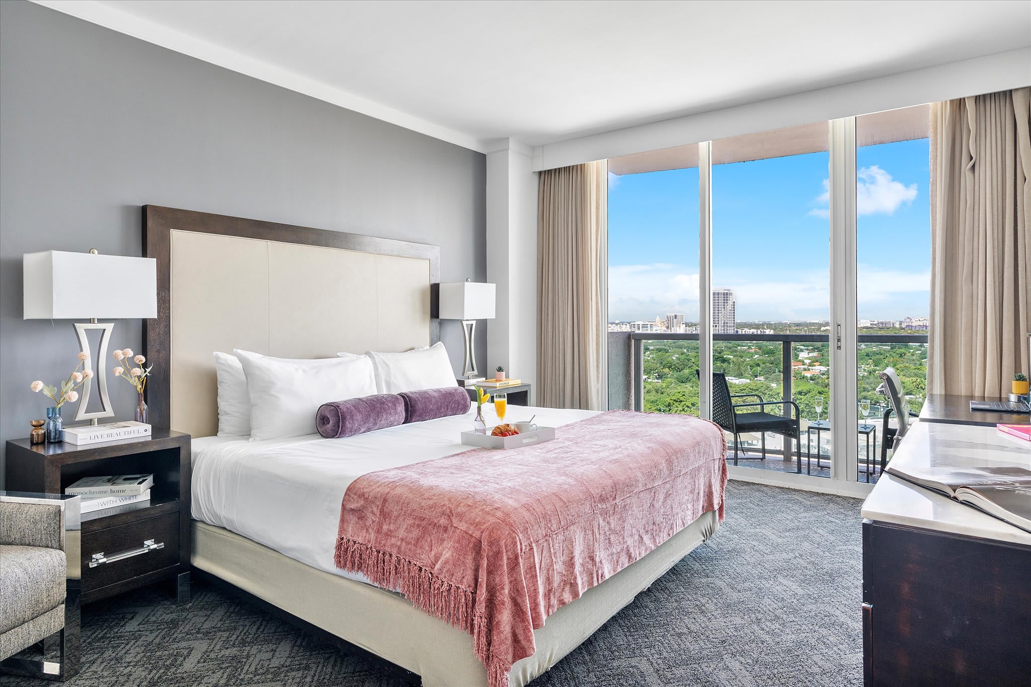 Spacious bedroom with a large bed featuring a tall upholstered headboard and a pink throw blanket. Two modern nightstands with lamps flank the bed, and floor-to-ceiling glass doors lead to a balcony with a city view.