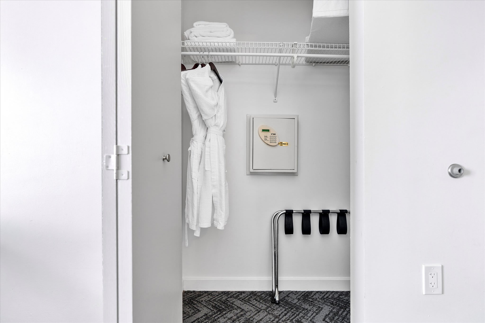 Walk-in closet with a white waffle-knit bathrobe hanging, wire shelving, a wall-mounted safe, and a luggage rack on a dark patterned carpet. The space is clean and organized, ready for guest use.