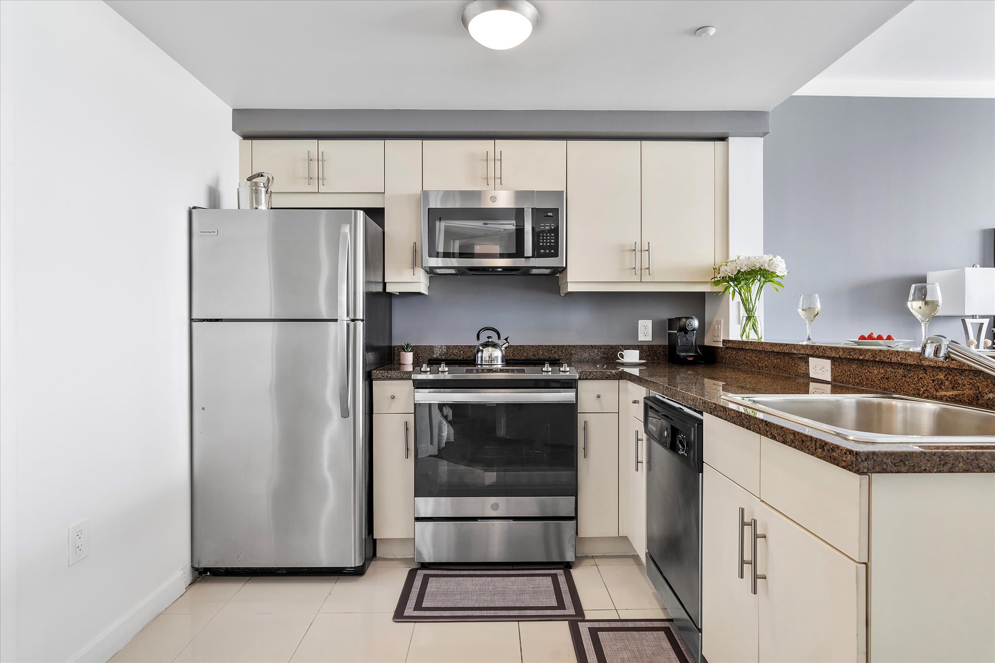 Full view of the modern kitchen with light beige cabinets and stainless steel appliances, including a large refrigerator, microwave, oven, and dishwasher. Dark granite countertops and a coffee maker are also visible.