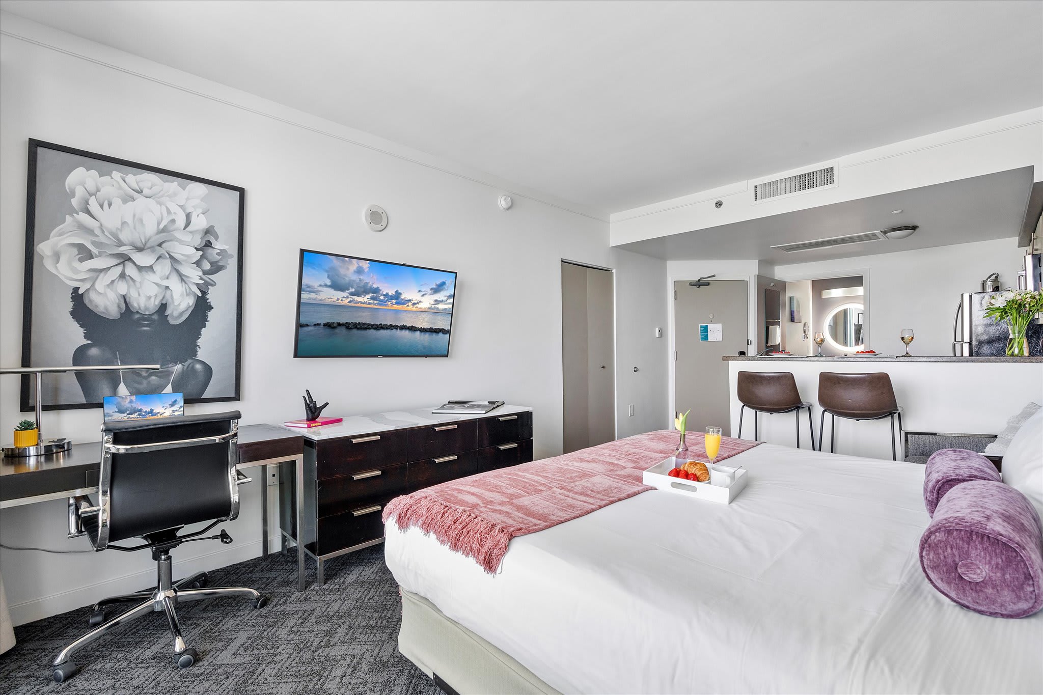 Open-concept studio apartment featuring a large bed with a pink throw, a dark wood desk with a black office chair, and a wall-mounted TV. A breakfast bar with two stools is visible in the background, connecting to the kitchen area.