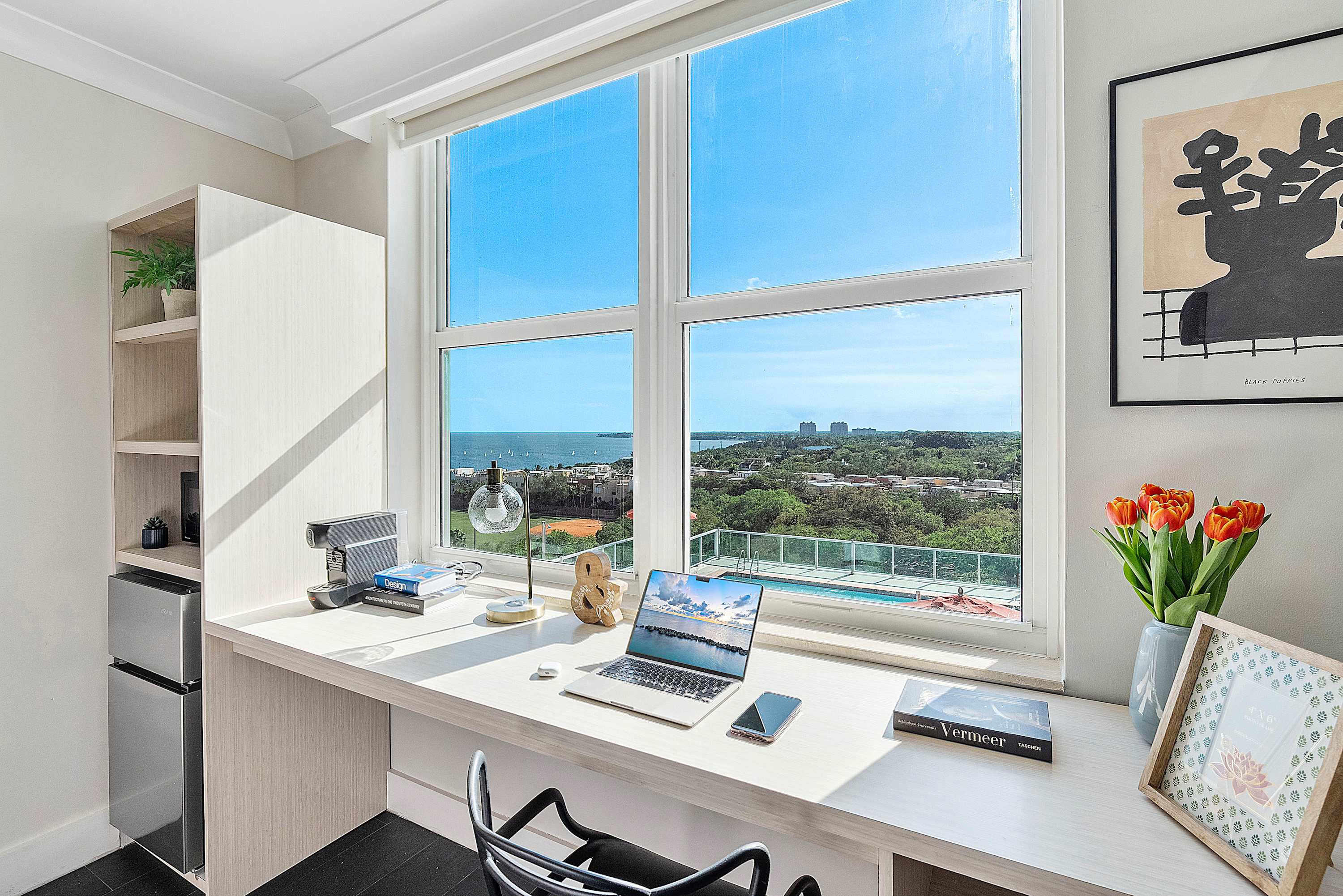 Dedicated workspace with a light wood desk, laptop, coffee maker, and mini-fridge, positioned by a large window offering expansive views of the bay and a rooftop pool. Shelving provides additional storage.