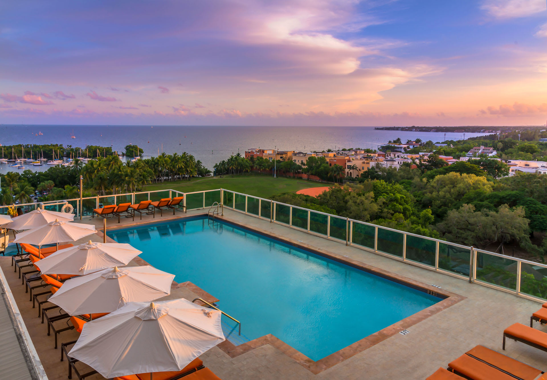Elevated outdoor swimming pool with lounge chairs and umbrellas, offering panoramic views of a bay filled with boats and a distant cityscape under a vibrant sunset sky. The pool area is surrounded by a glass railing.