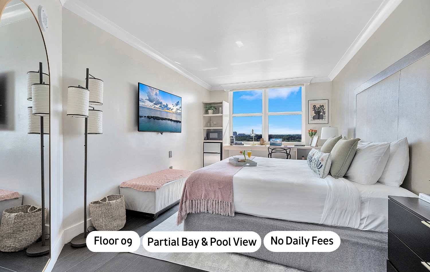 Bright bedroom featuring a large bed with white and pink bedding, a wall-mounted TV, and a desk area by a window offering partial bay and pool views. The room has light walls, dark flooring, and a modern aesthetic.