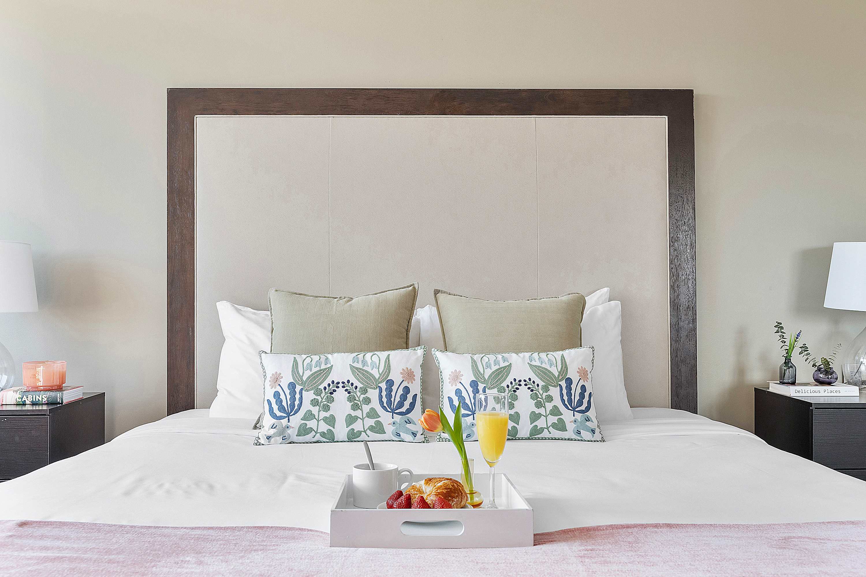 Close-up view of a comfortable bed featuring a stylish dark wood and upholstered headboard, crisp white linens, and decorative pillows. A breakfast tray with fruit and juice is placed on the bed.