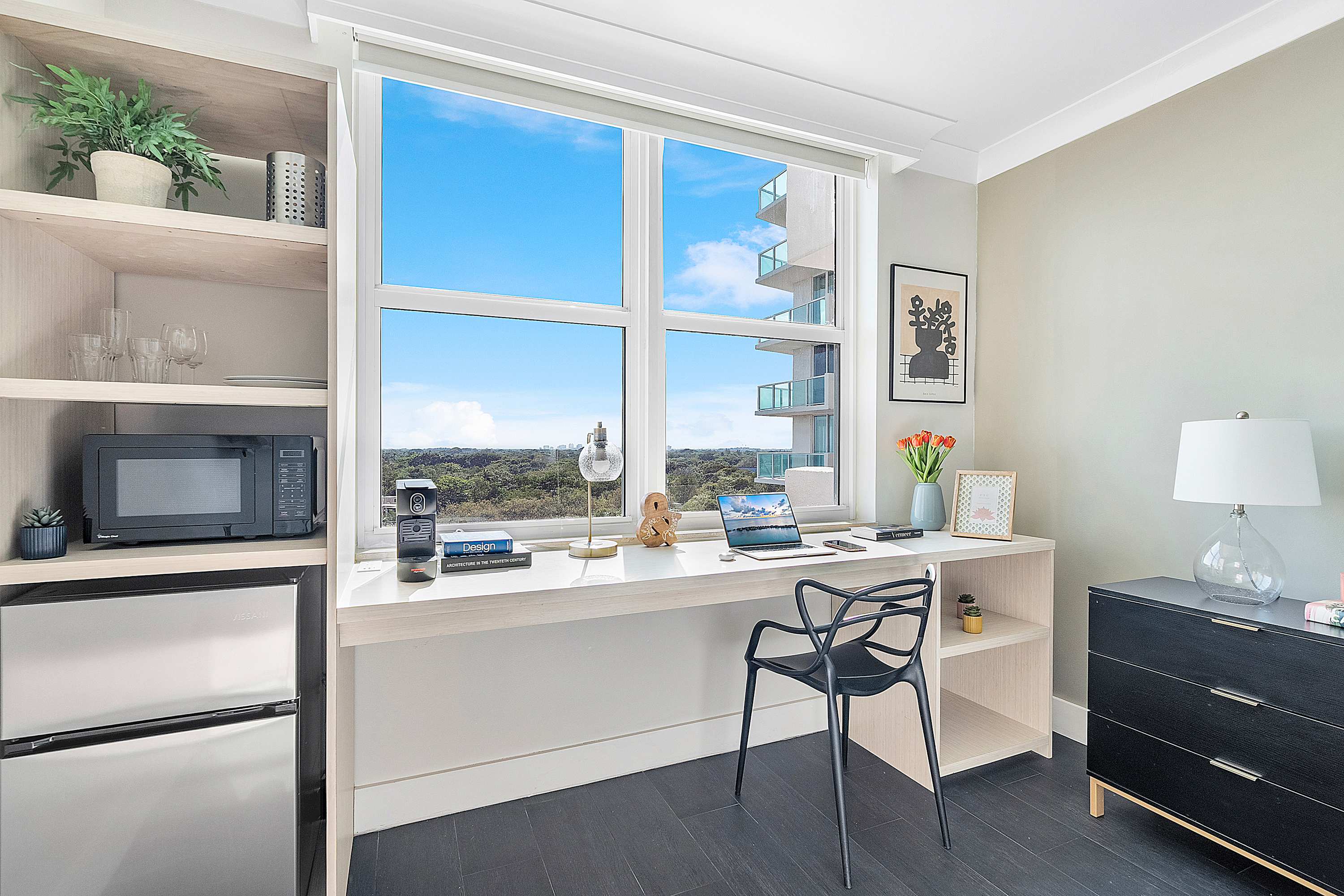 Well-equipped workspace featuring a light wood desk with a laptop, microwave, and mini-fridge, set against a large window offering bright views of the surrounding area and distant water.