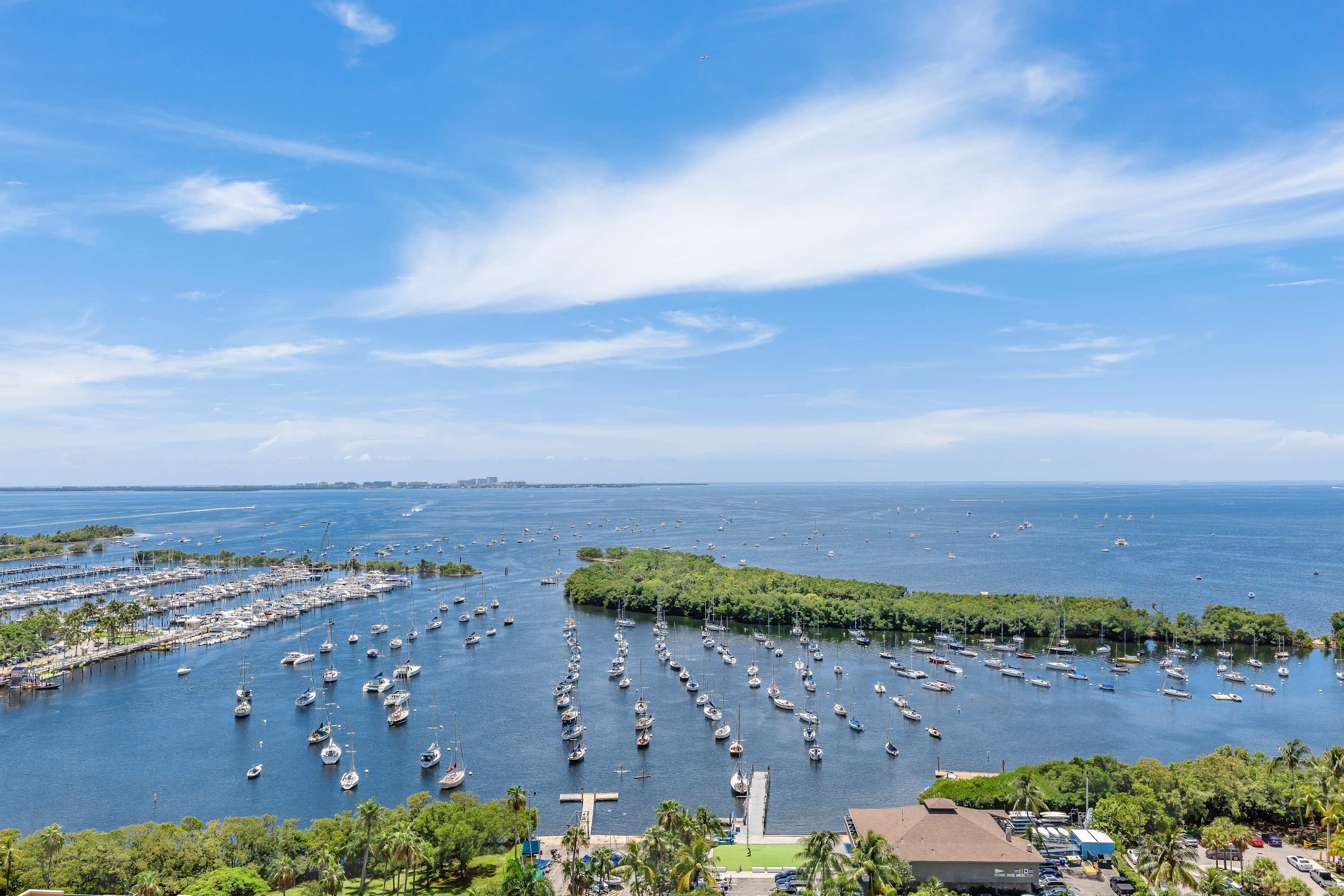 A high-angle view captures a vast expanse of blue water dotted with numerous sailboats and a bustling marina, with a lush green island and distant city skyline under a bright, cloudy sky.