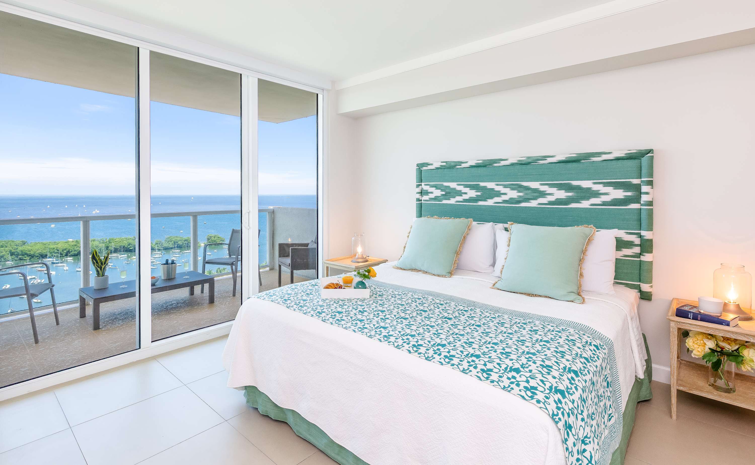 Bright bedroom with a queen-sized bed featuring a teal and white patterned headboard and bedding, light tile flooring, and floor-to-ceiling sliding glass doors leading to a private balcony with bay views.