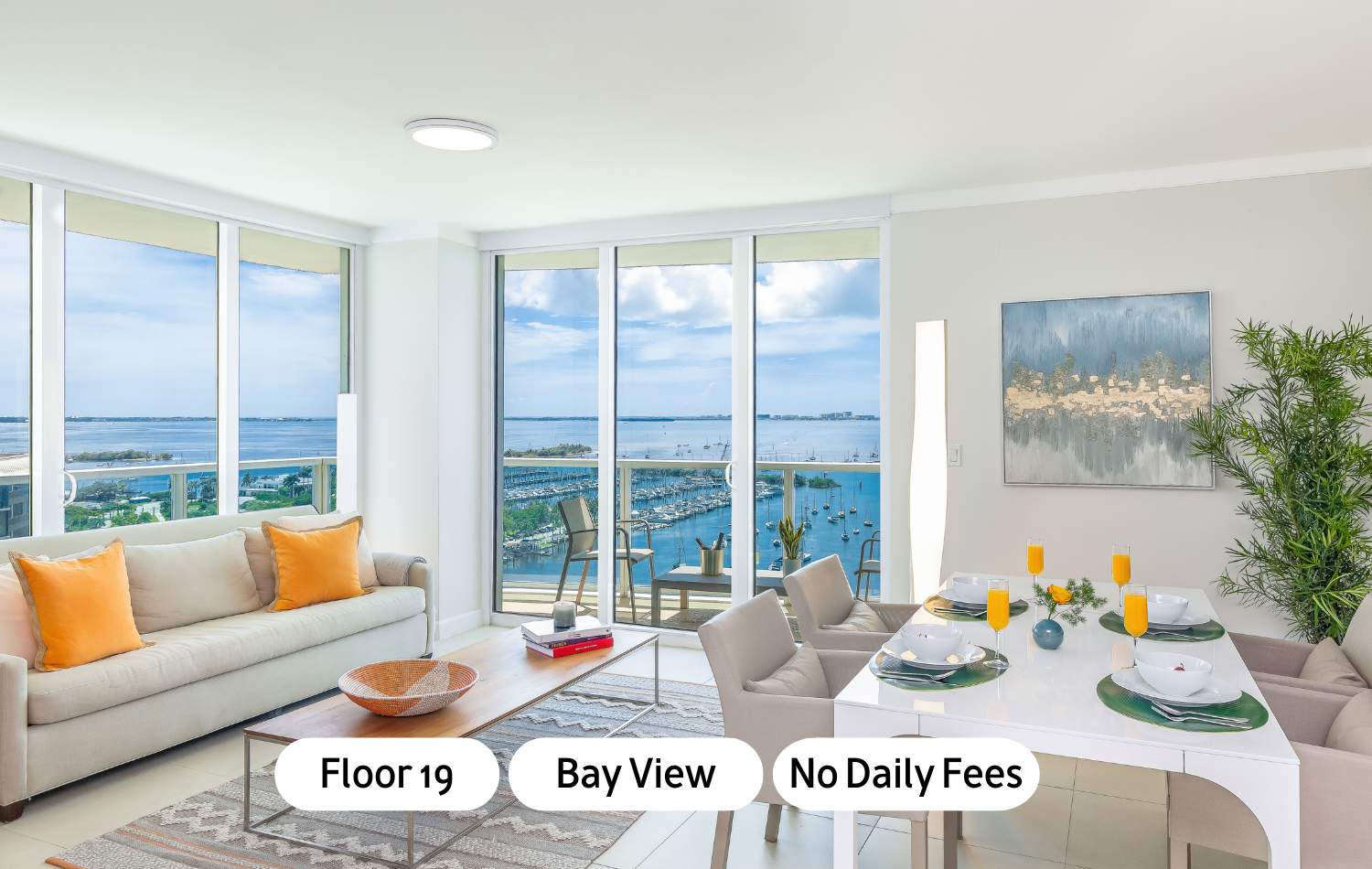 Bright, open-concept living and dining area with white walls, light tile flooring, a beige sofa with orange pillows, a wooden coffee table, and a white dining table set for six, all overlooking a bay with boats through large windows and a sliding glass door to a balcony.