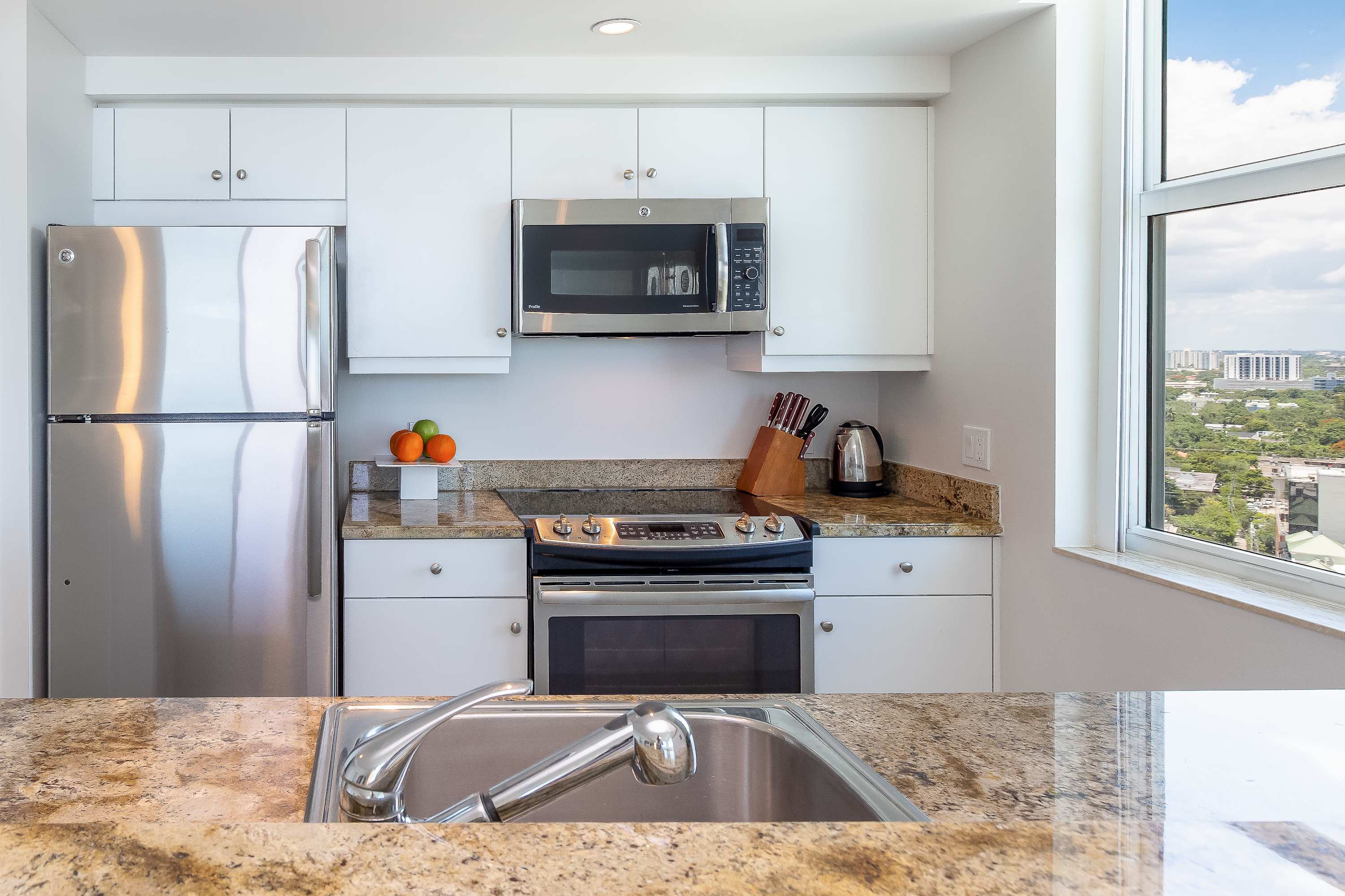 Compact, modern kitchen with white cabinetry, granite countertops, stainless steel appliances including a refrigerator, microwave, and electric range, and a window offering a city view.