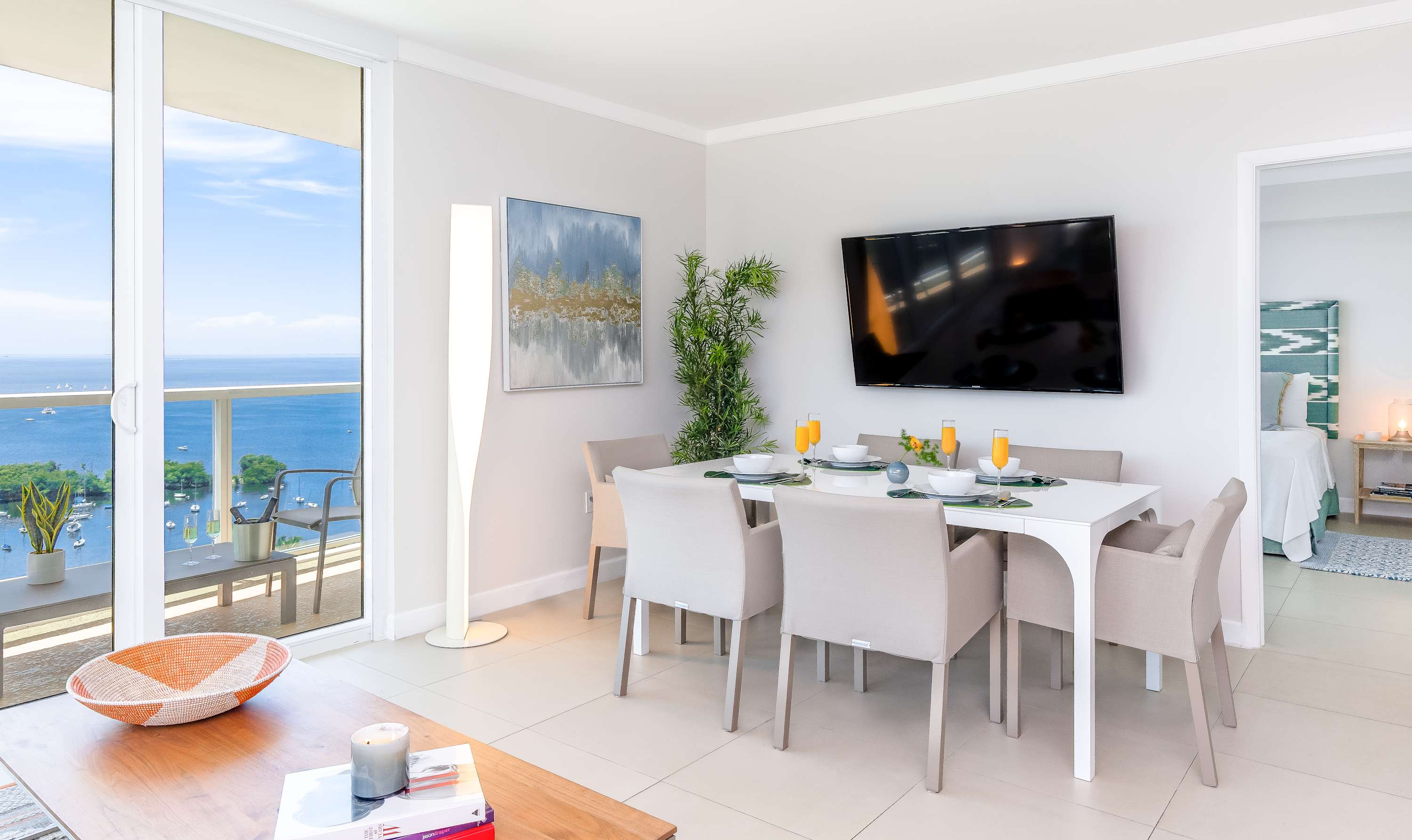 Contemporary dining area with a white rectangular table set for six, light grey upholstered chairs, a tall floor lamp, and a wall-mounted TV, adjacent to a living area and offering bay views through large windows.