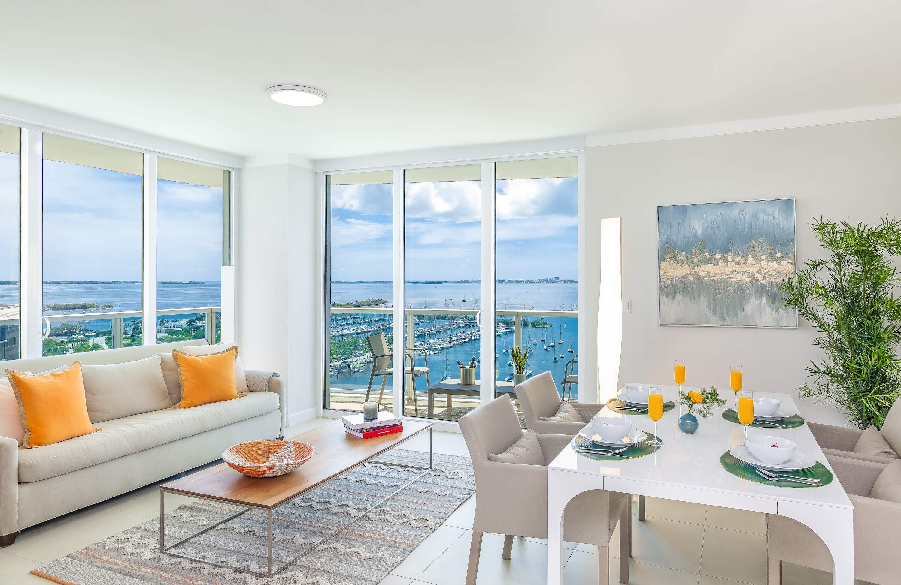 Modern, open-concept living and dining space with white walls, light tile flooring, a beige sofa with orange throw pillows, a wooden coffee table, and a white dining table with six chairs, all offering expansive bay views through floor-to-ceiling windows and a balcony door.