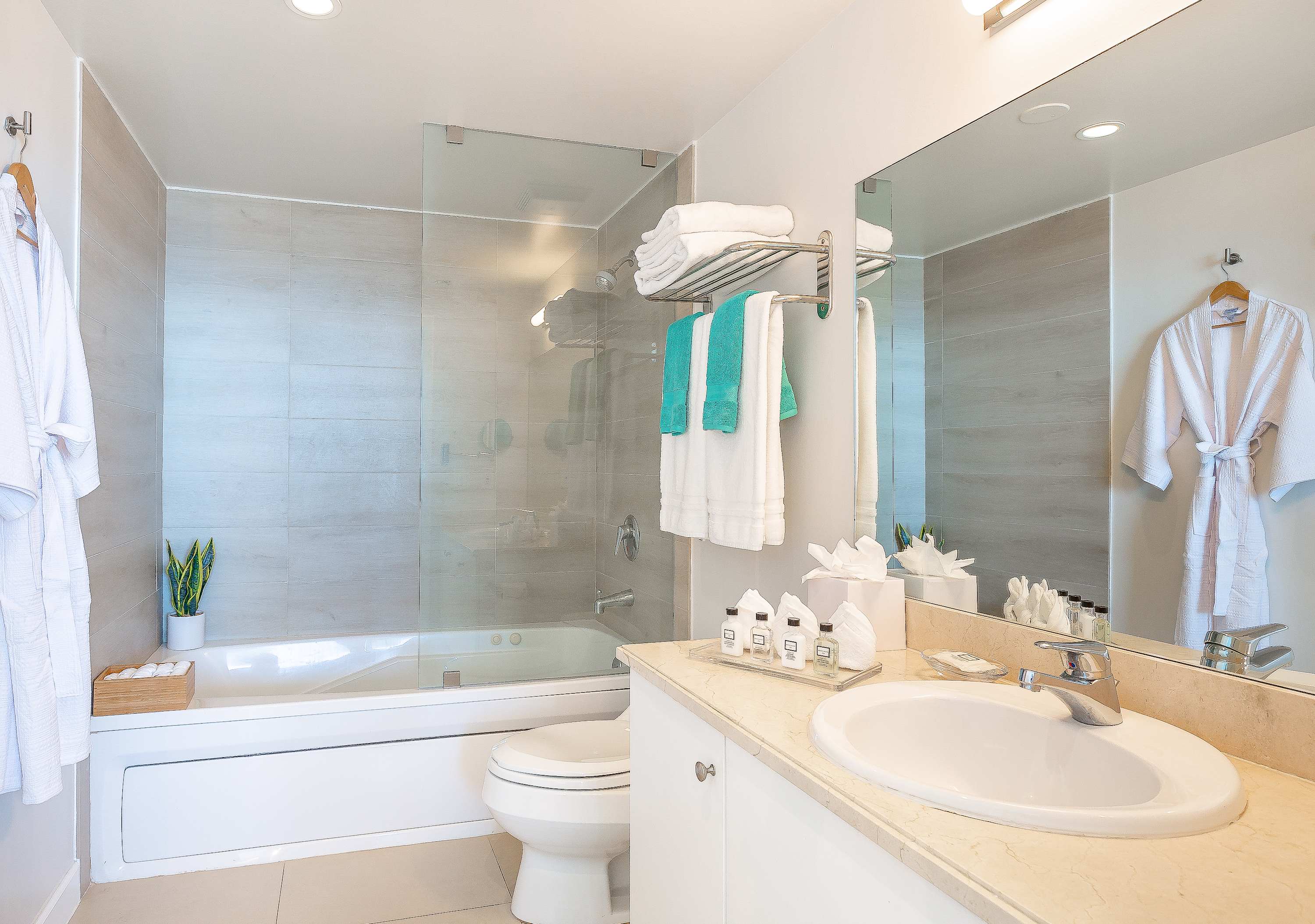 Clean, modern bathroom with a single vanity, light-colored countertop, large mirror, and a combination shower/bathtub with a glass enclosure, featuring light grey tiled walls and white bathrobes.