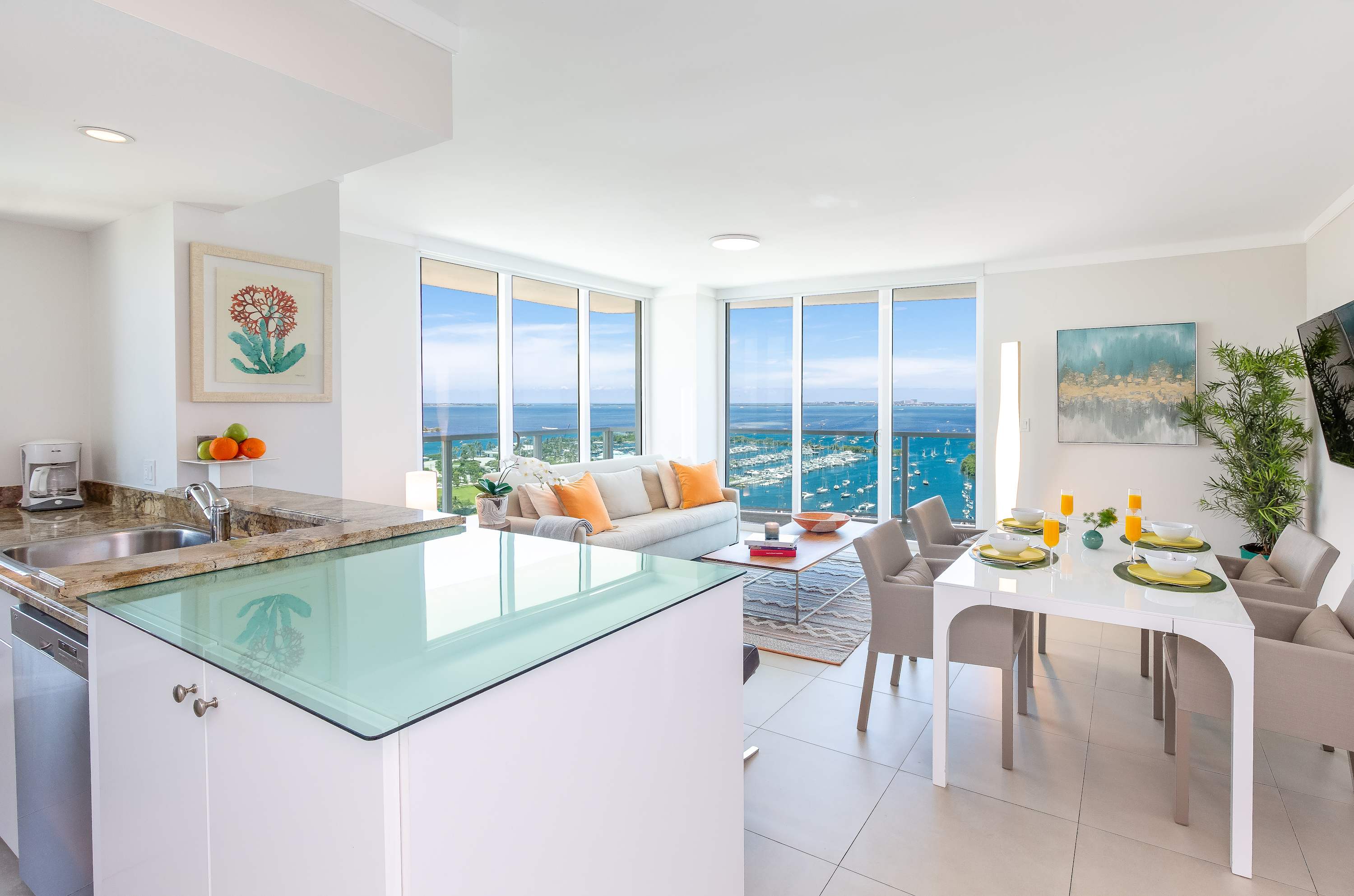 Open-concept living space combining a modern kitchen with white cabinets and granite countertops, a dining area with a white table, and a living room with a beige sofa, all featuring light tile flooring and panoramic bay views through extensive windows.