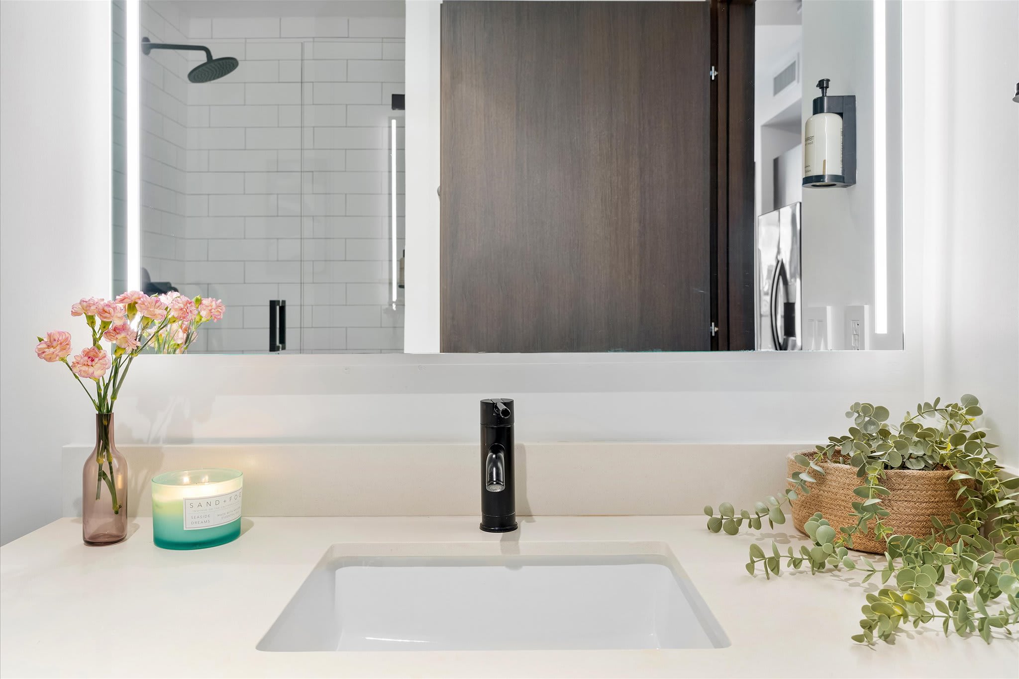 A close-up of the bathroom vanity with a white countertop, integrated sink, and a sleek black faucet. An illuminated mirror reflects the shower, and decorative plants and a candle add a touch of warmth.