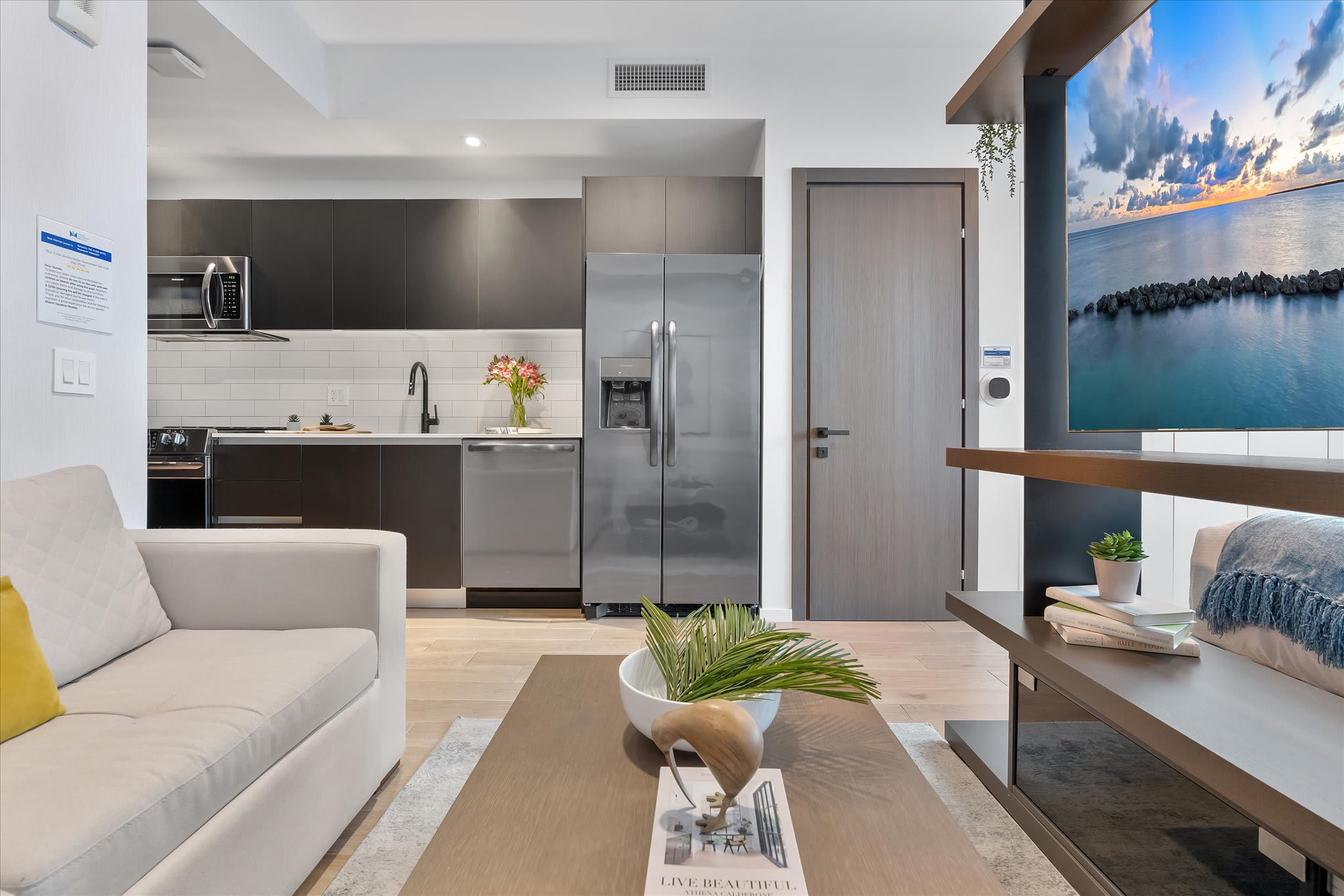 A modern kitchen with dark flat-panel cabinetry, white subway tile backsplash, and stainless steel appliances, including a refrigerator and dishwasher. A light-colored sofa and coffee table are visible in the foreground.
