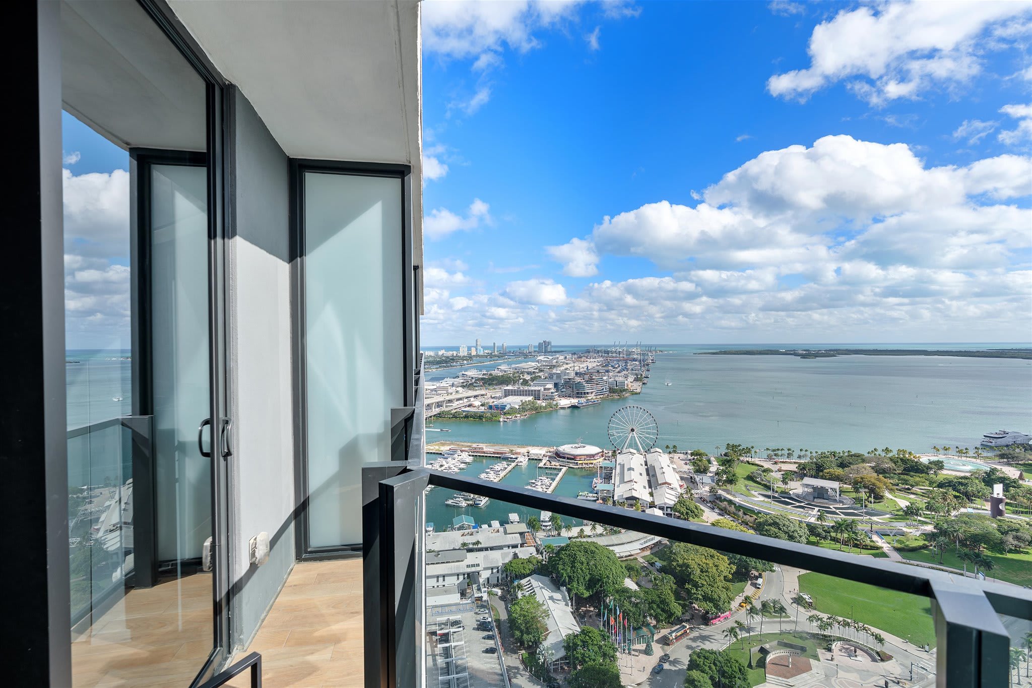 Private balcony with a black railing and glass doors, offering an expansive panoramic view of the city skyline, a bustling marina with boats, and the vast bay under a bright blue sky with white clouds.