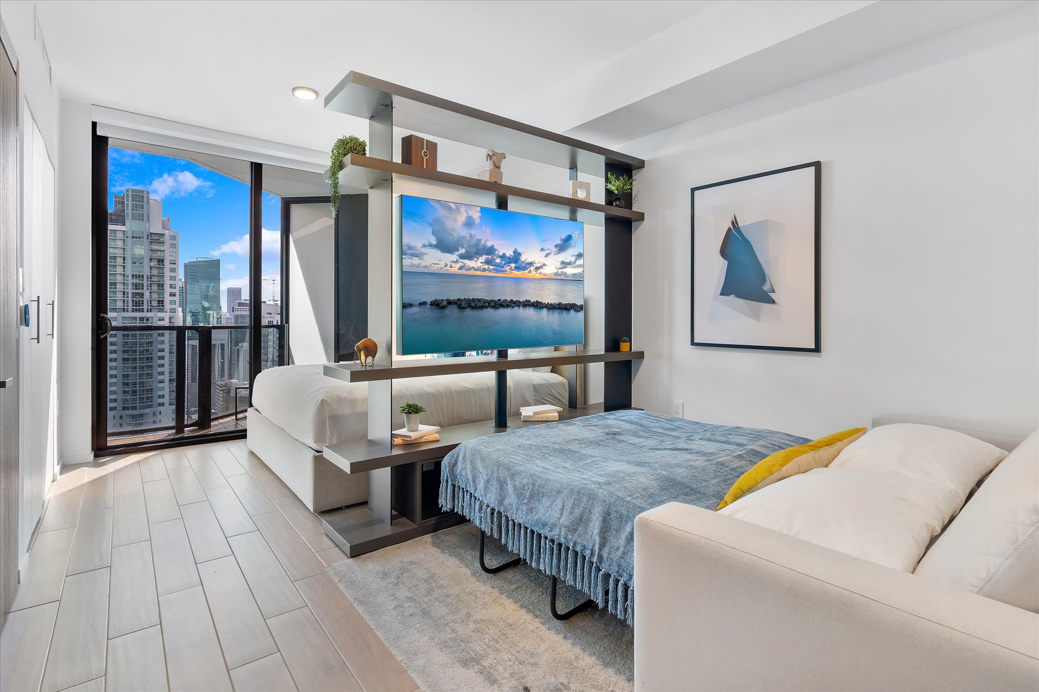 A modern studio living space with a sofa bed, a large TV on a room divider, and a balcony offering panoramic city views. Light wood-look flooring and white walls create a bright atmosphere.
