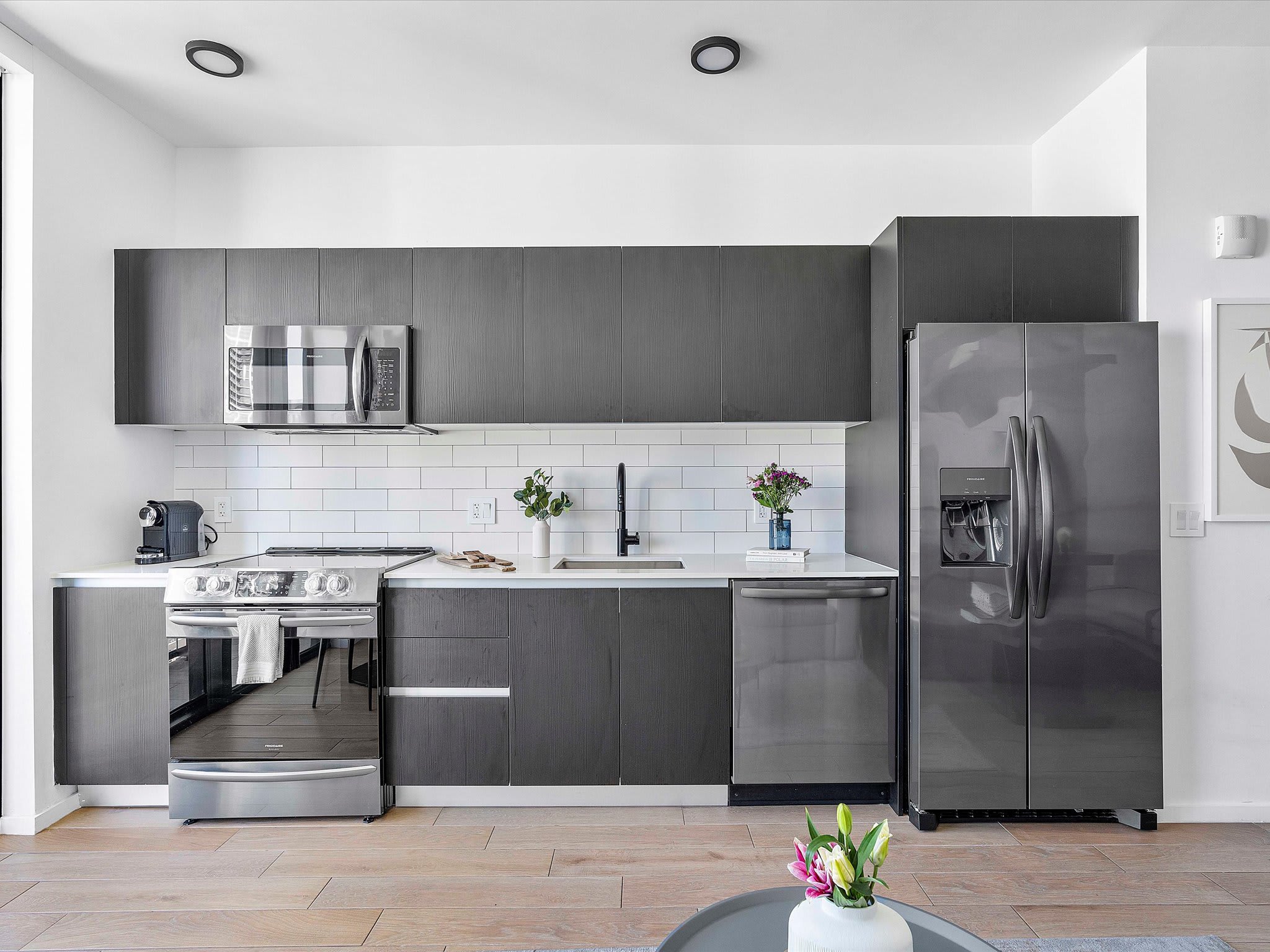 Modern kitchen featuring dark gray flat-panel cabinetry, white quartz countertops, and a white subway tile backsplash. Stainless steel appliances include a microwave, range, dishwasher, and a large French-door refrigerator.