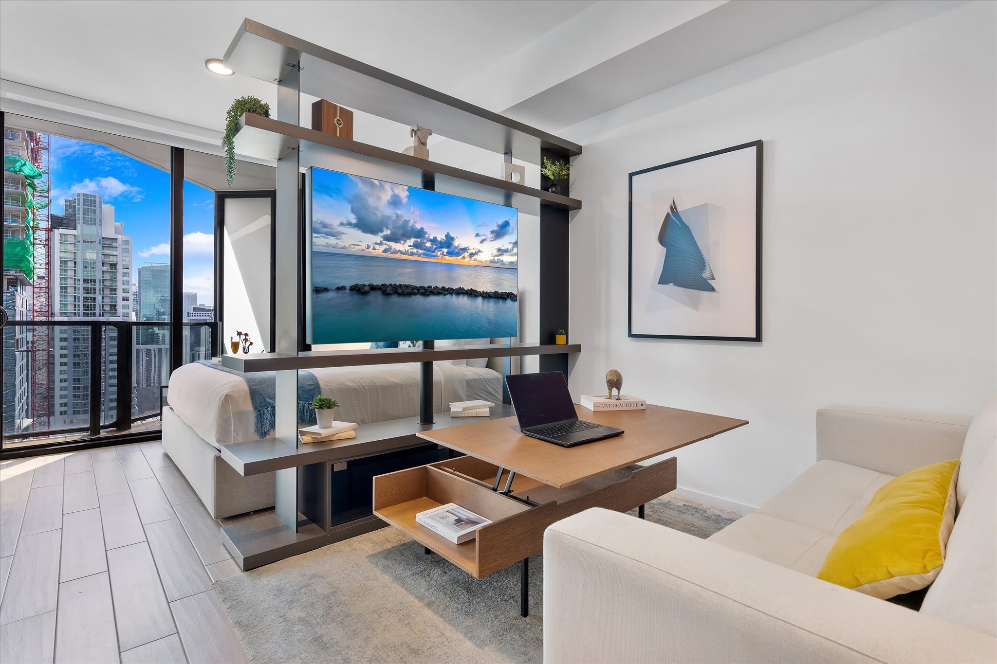Modern living space featuring a light beige sofa, a stylish wooden coffee table with storage, and a large flat-screen TV integrated into a dark shelving unit. Floor-to-ceiling windows offer a glimpse of the city's high-rise buildings and construction.