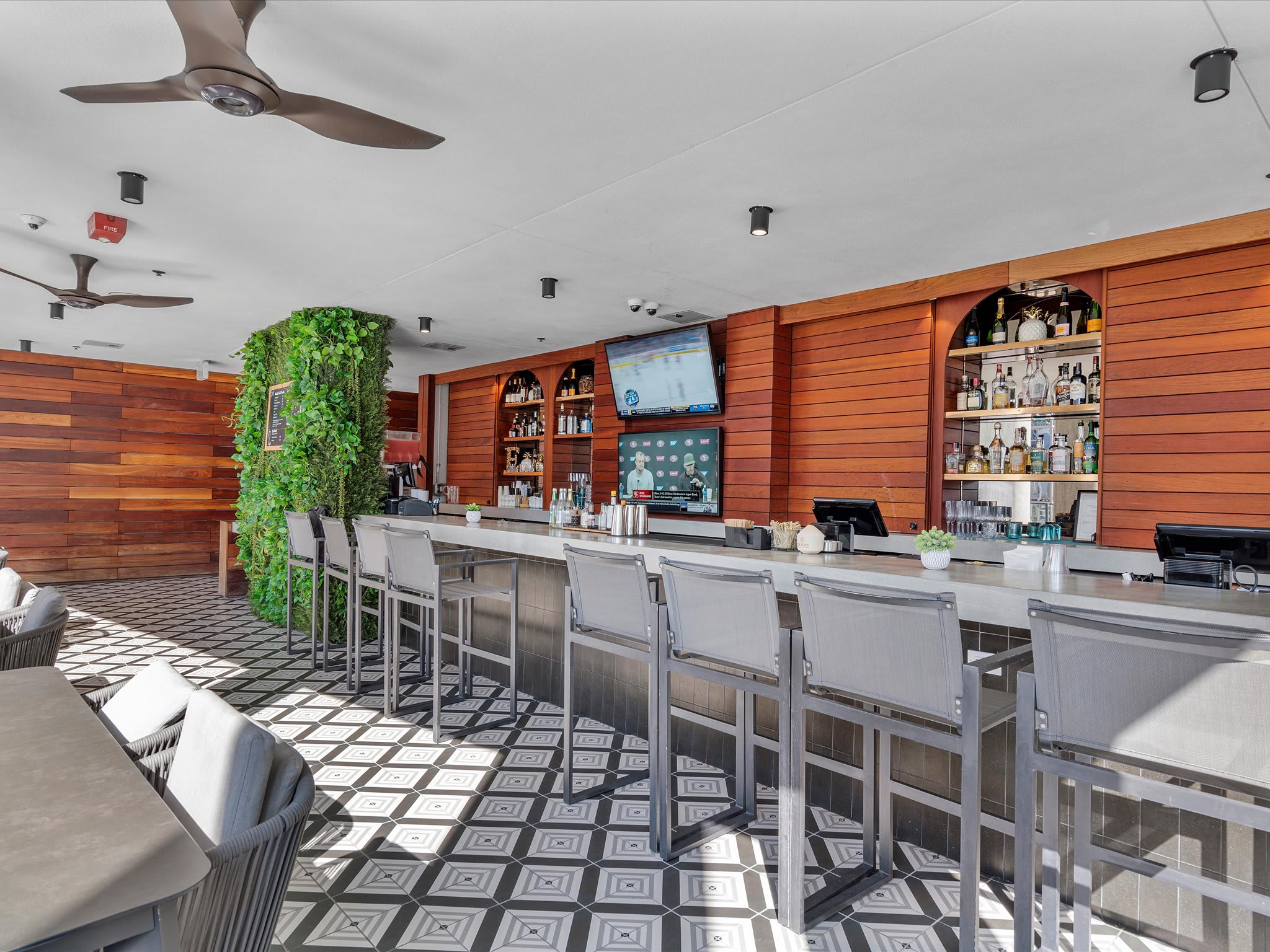 A stylish bar area with a long white countertop, high stools, and wooden paneling on the walls. Shelves filled with bottles are visible behind the bar, and a large TV screen is mounted above.
