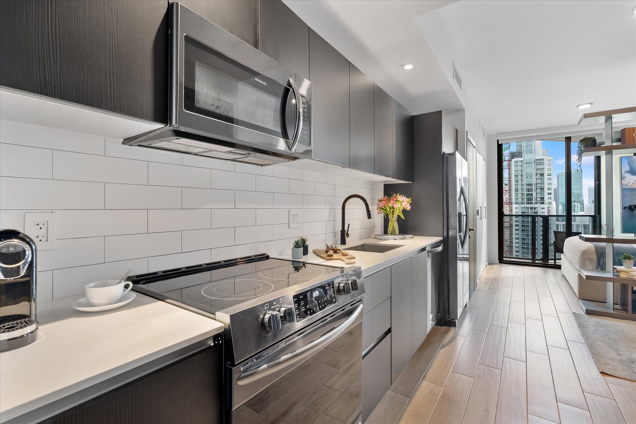 A detailed view of the modern kitchen featuring a stainless steel electric range, microwave, and sink with a black faucet. White subway tile backsplash and light countertops complement the dark cabinetry.