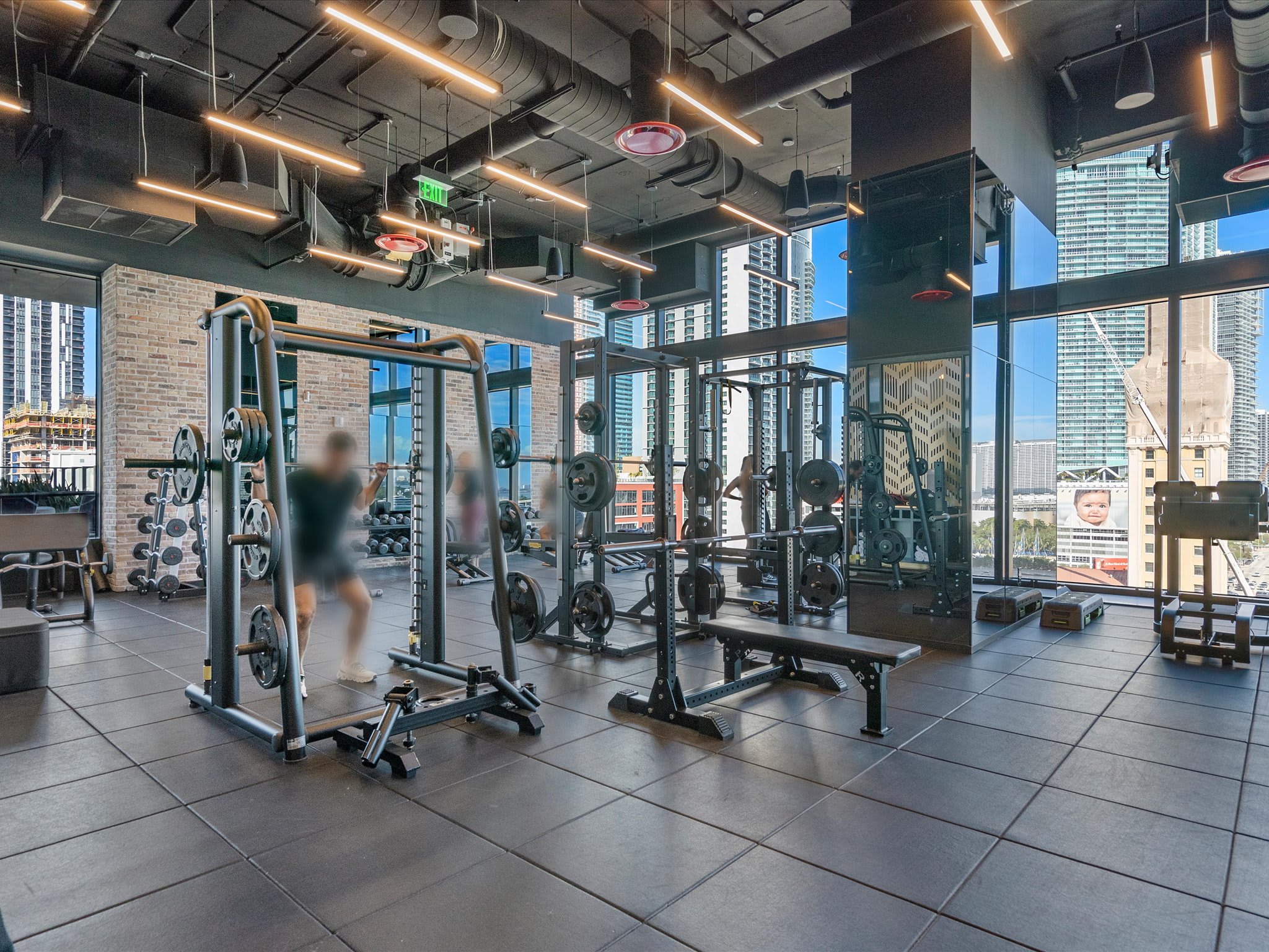A modern fitness center with exposed black ceilings, industrial lighting, and large windows offering city views. It features various weightlifting equipment, including squat racks and benches, on a dark tiled floor.