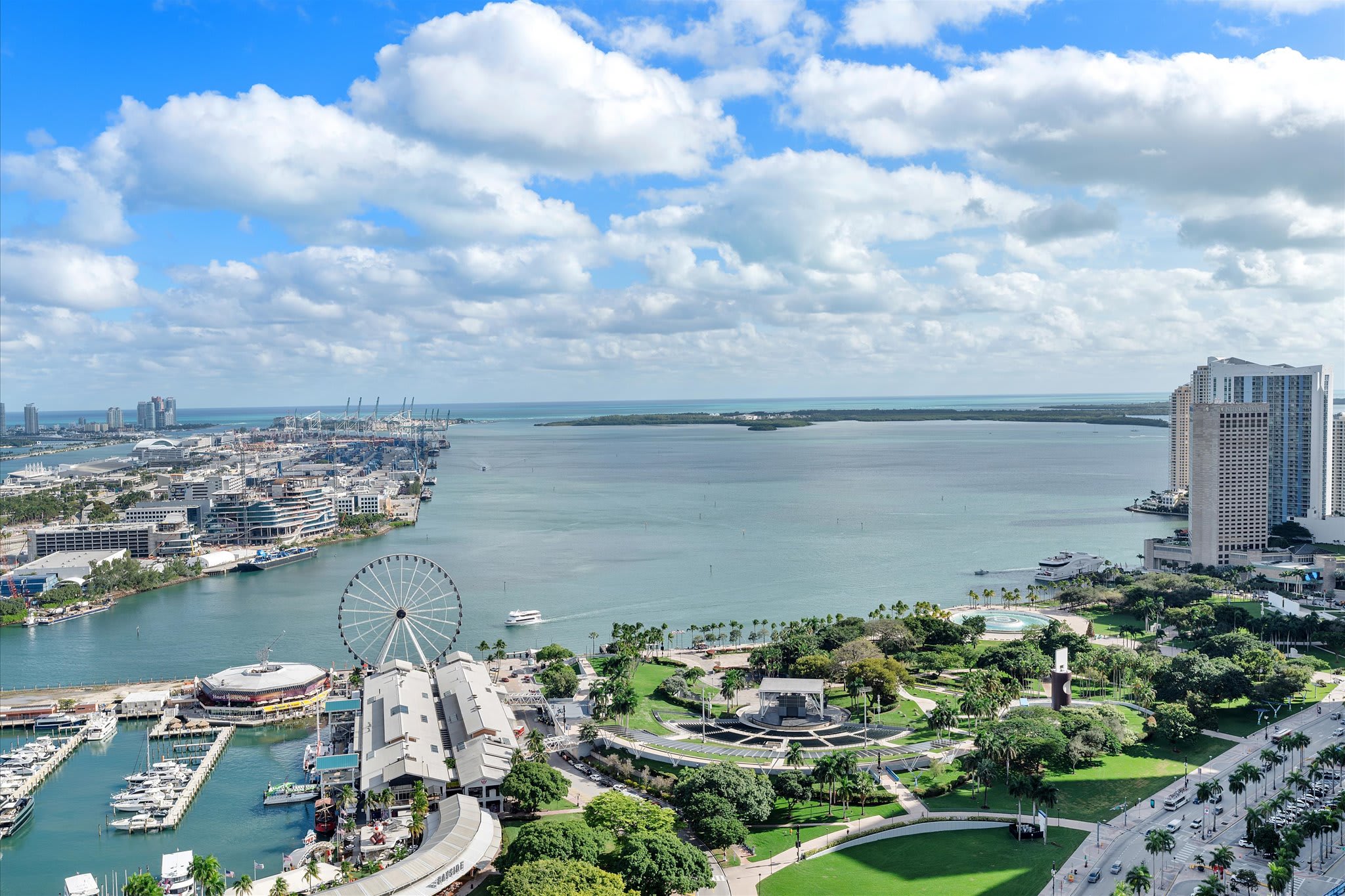 Panoramic aerial shot capturing the expansive Miami bay area, featuring a busy port with cargo ships, a large Ferris wheel, a marina, and lush green parks. The city skyline stretches along the distant coastline under a bright, cloud-dotted sky.