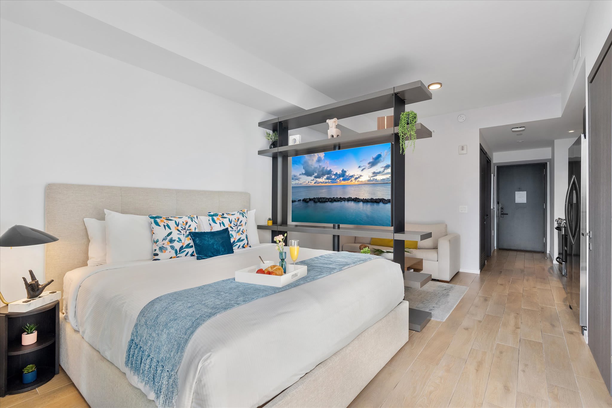 The studio's sleeping area features a large bed with a light headboard and colorful pillows, facing a TV mounted on a stylish room divider. A breakfast tray with drinks is placed on the bed.