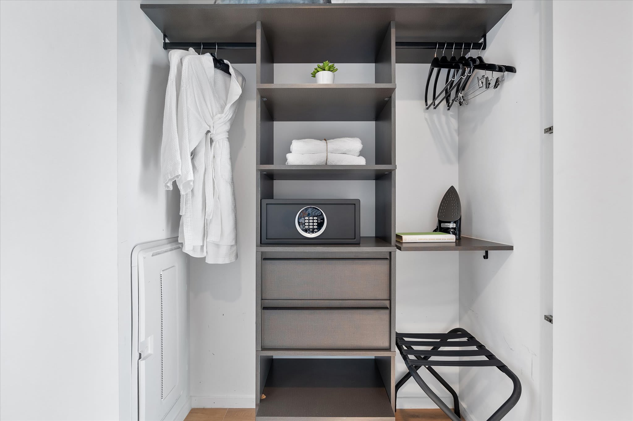 A well-organized closet with shelving, hanging space, and drawers, featuring a safe, an iron, and a luggage rack. White robes and fresh towels are neatly arranged.