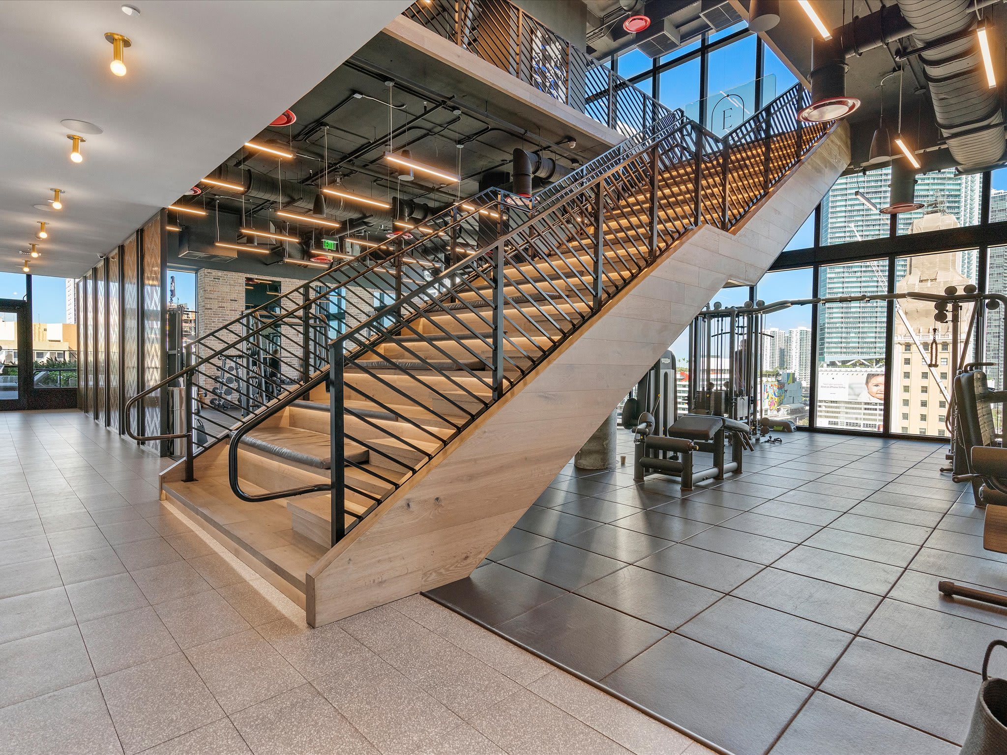 A grand, modern wooden staircase with black metal railings connects multiple levels in a bright, open space. Large windows offer city views, and gym equipment is visible on the lower level.