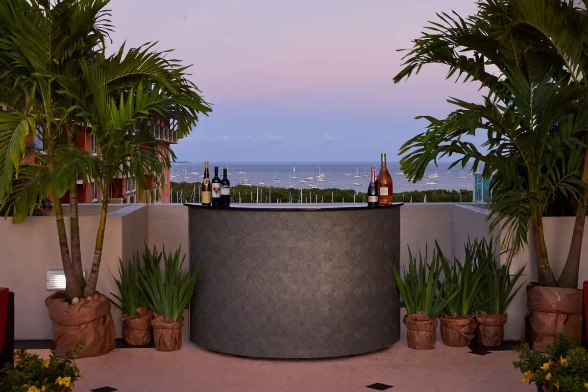 An elegant outdoor bar area featuring a dark, textured bar counter flanked by potted palm trees and lush green plants, set against a twilight backdrop of a bay dotted with boats. Bottles of wine are displayed on the bar.