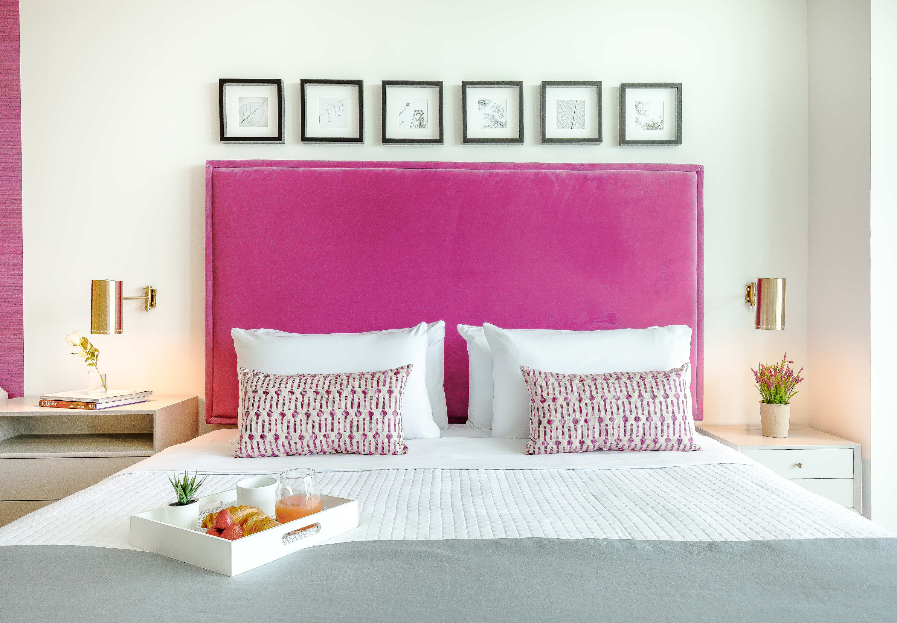 A comfortable king-sized bed with a plush hot pink headboard, adorned with white and patterned pink pillows. Two modern nightstands with wall-mounted lamps flank the bed.