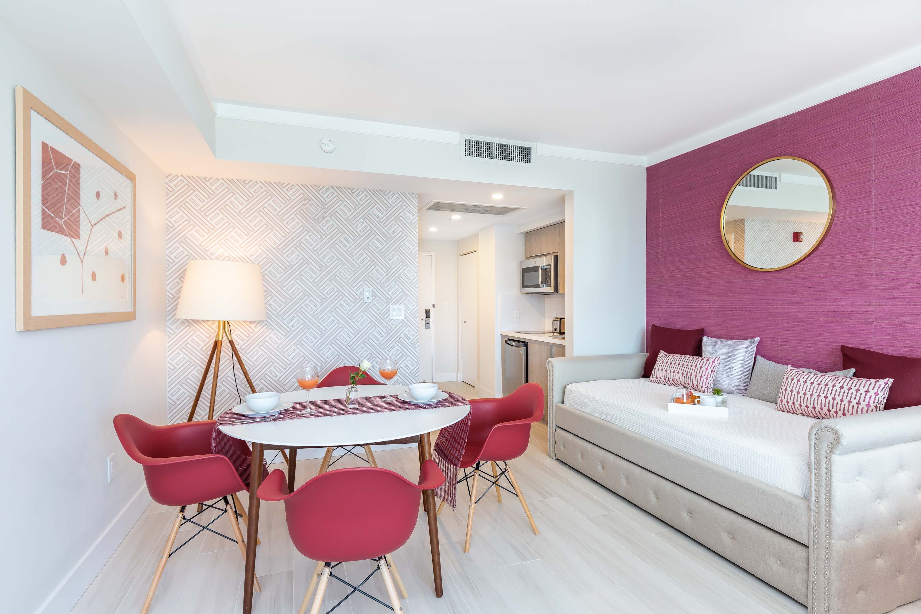 A bright studio space featuring a round white dining table with four red chairs, a comfortable daybed, and a fuchsia accent wall with a round mirror.