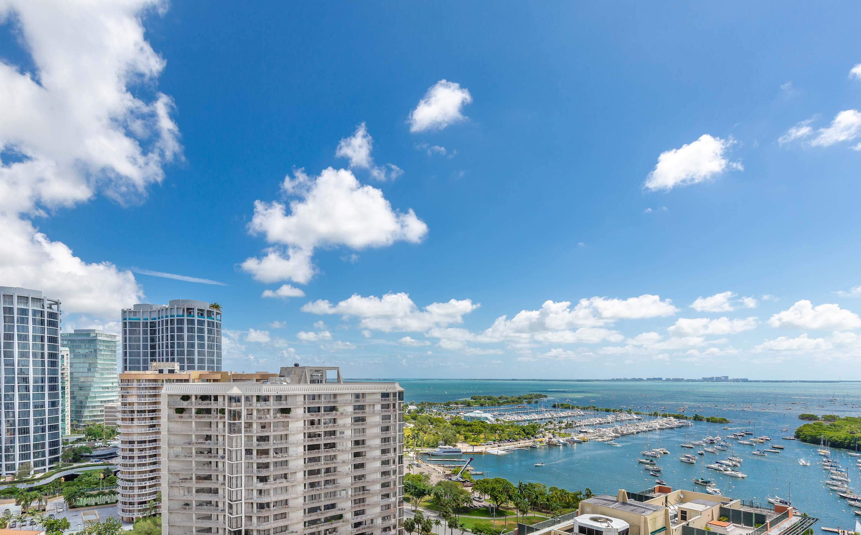 A panoramic view from a high-rise balcony, showcasing a bustling marina filled with boats and the expansive bay, framed by other modern high-rise buildings under a bright blue sky with scattered clouds. The water is a vibrant turquoise.