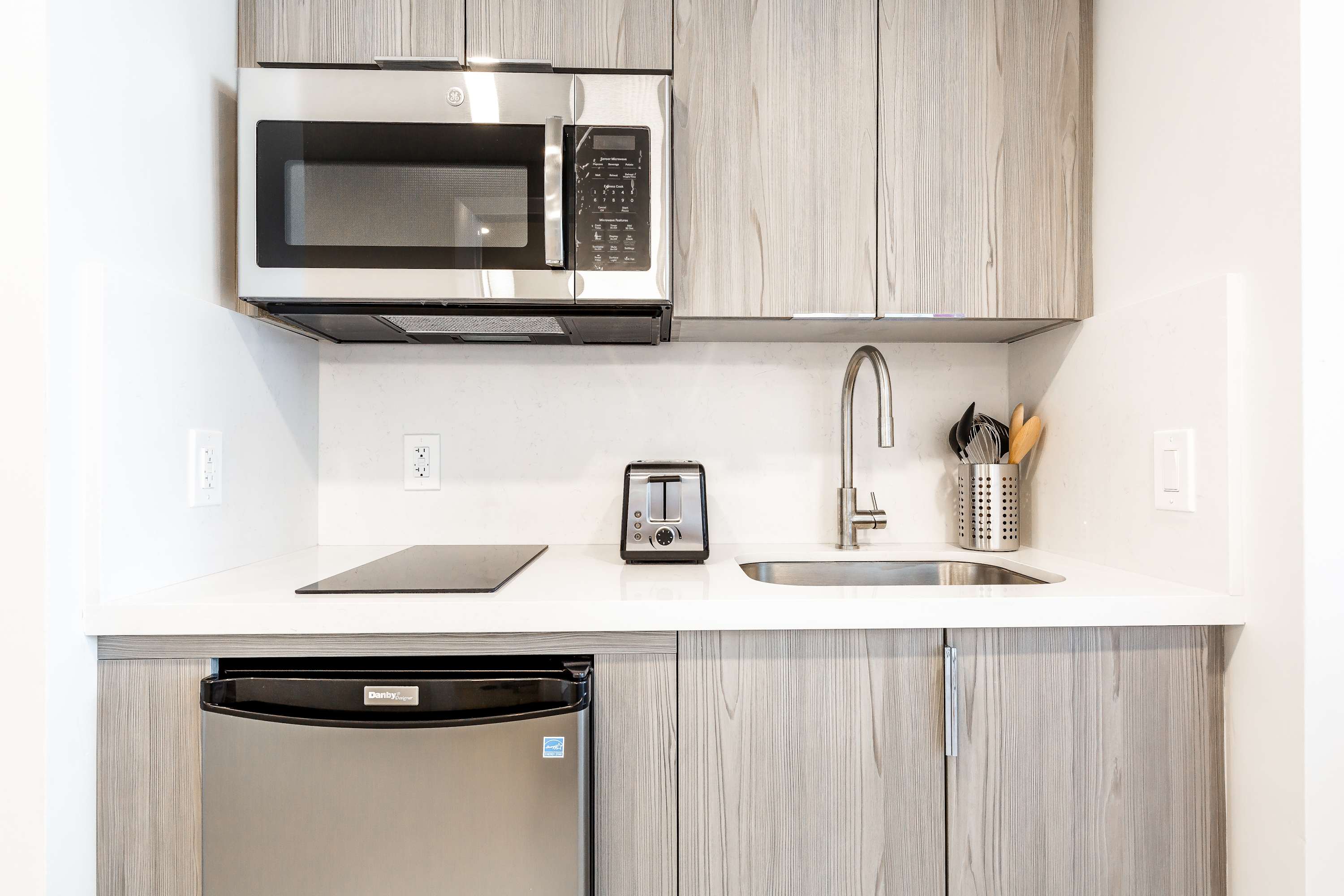 A compact and modern kitchenette featuring light wood cabinets, white quartz countertops, a stainless steel microwave, a small refrigerator, and a toaster.