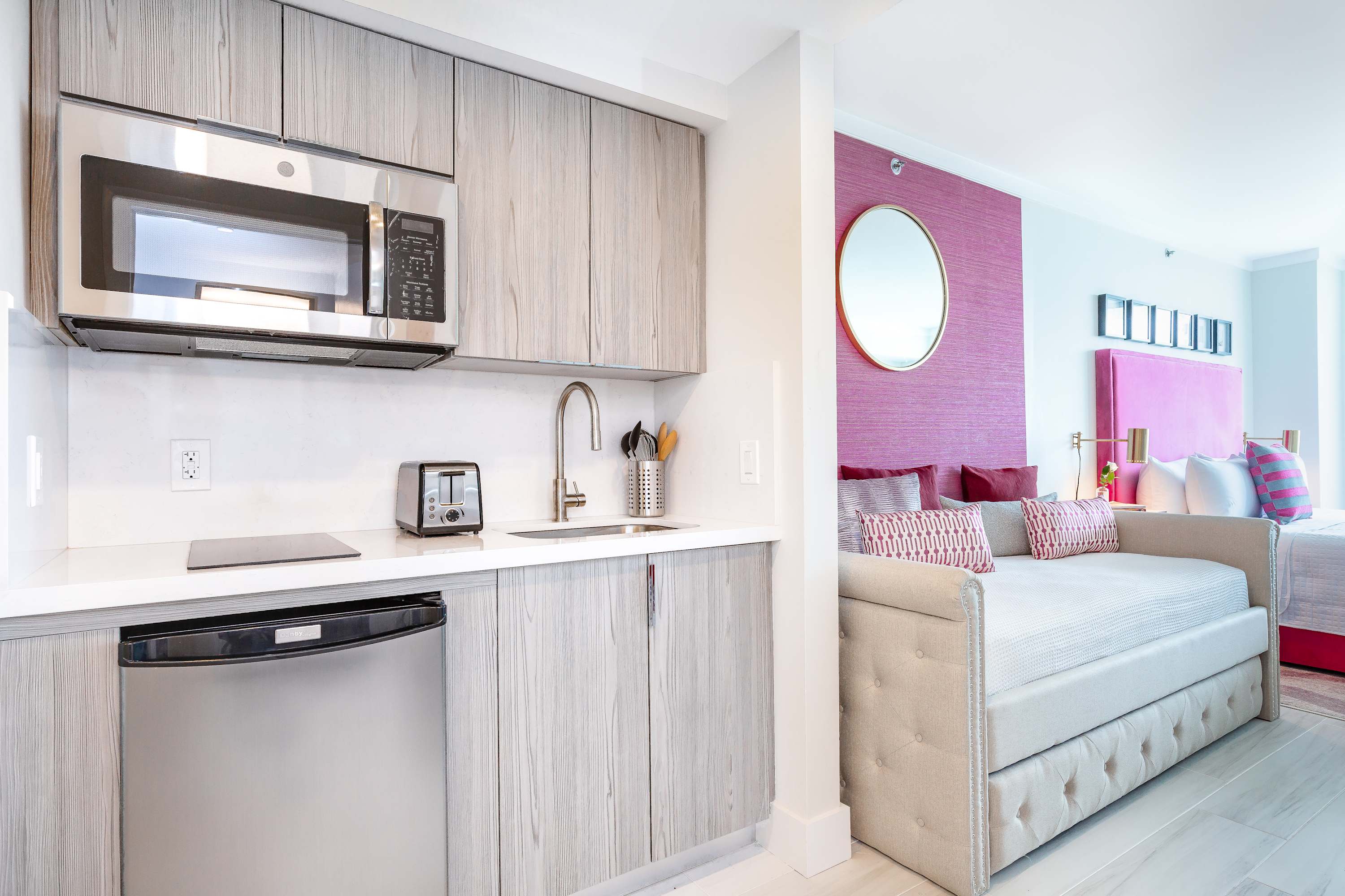 A well-appointed kitchenette with light wood cabinetry and stainless steel appliances, positioned adjacent to a comfortable daybed with decorative pillows, part of an open-plan studio.