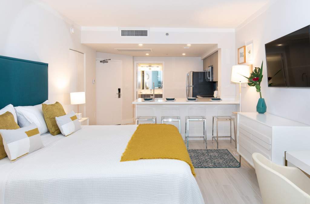 A wide shot of the studio apartment, showing the bed with a teal headboard and yellow accents, a white dresser with a TV, and a view towards the kitchen and bathroom entrance.