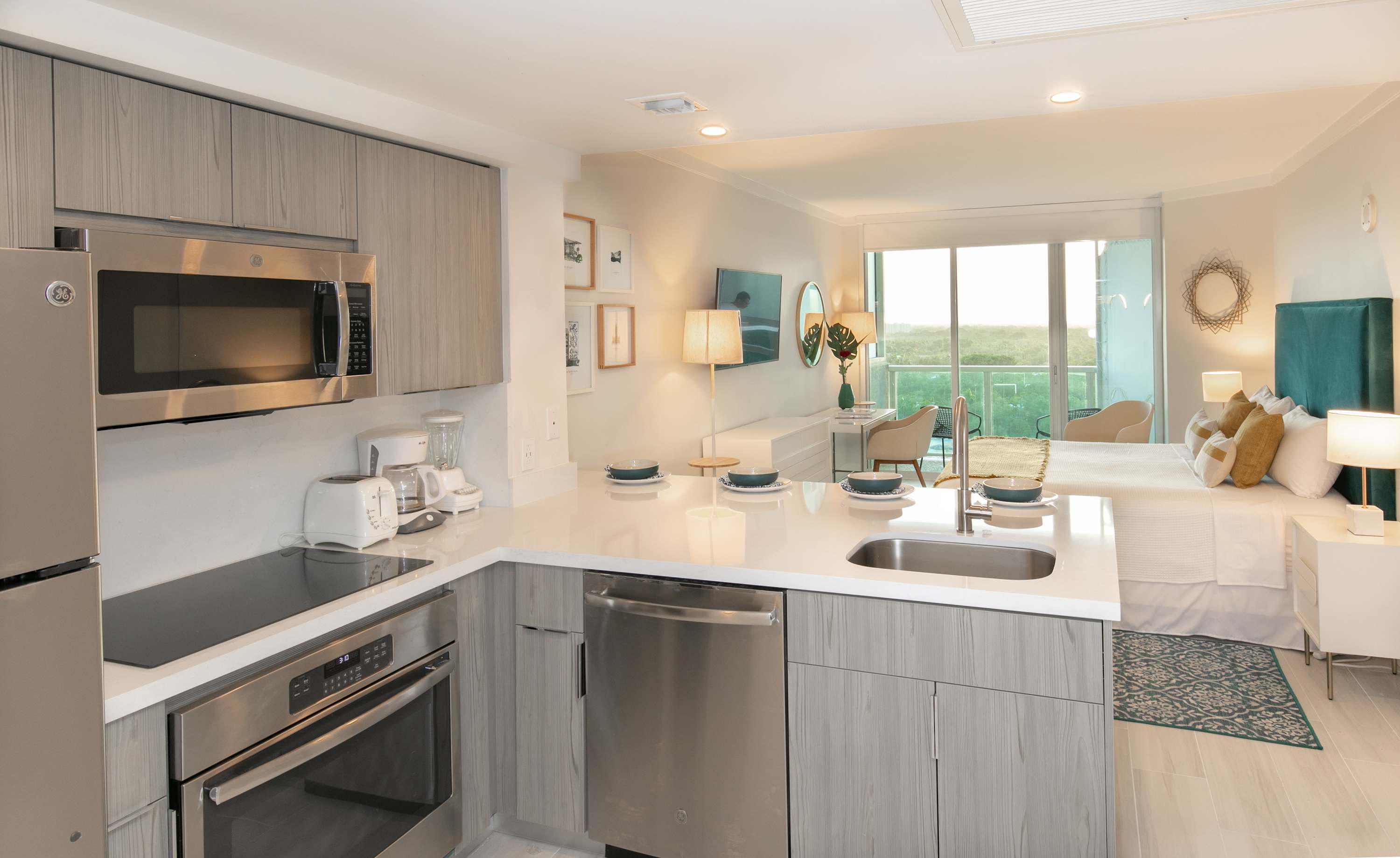 An open-concept view of the modern kitchen with light wood-grain cabinetry and stainless steel appliances, seamlessly flowing into the studio bedroom area with a bed and balcony access.