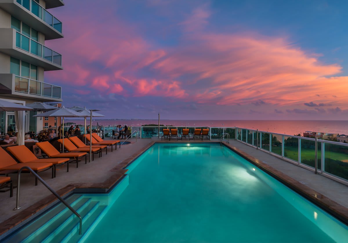 A stunning rooftop swimming pool area at sunset, featuring a rectangular pool surrounded by orange lounge chairs. The scene offers a breathtaking ocean view with a vibrant, colorful sky and a modern building in the background.