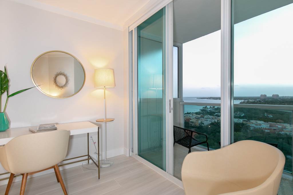 A corner of the bedroom featuring a white desk with two chairs, a round gold-framed mirror, a floor lamp, and large sliding glass doors opening to a balcony with expansive bay views.
