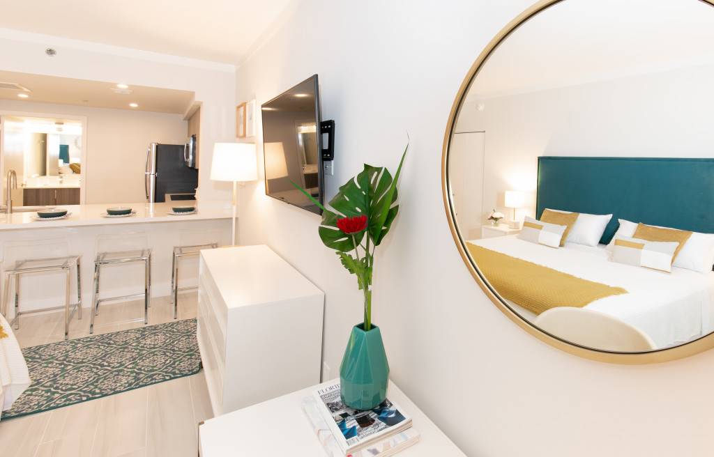 A view from the bed in the studio apartment, showing a large round mirror reflecting the bed, a white dresser with a plant, and the kitchen area with bar stools in the background.