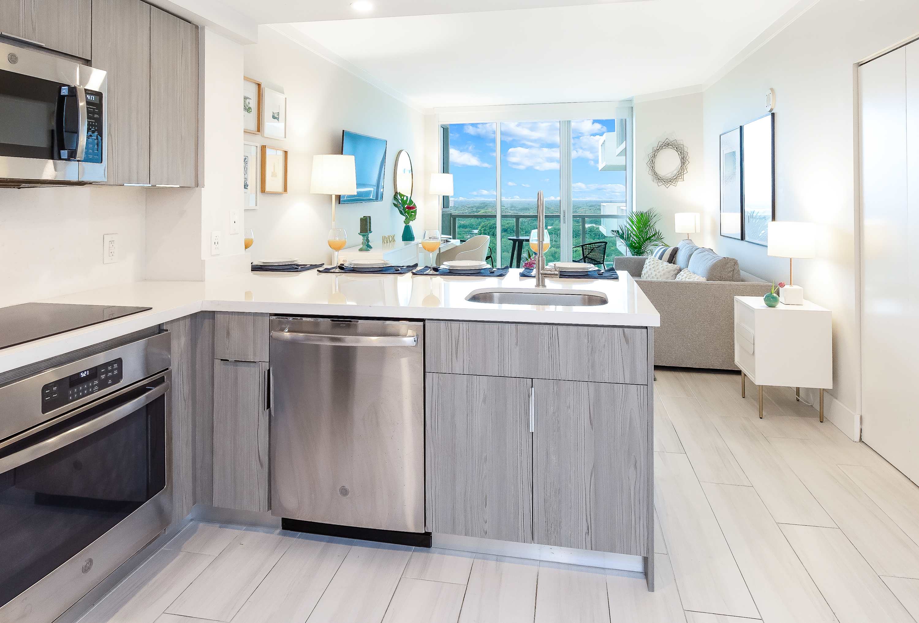 Modern kitchen featuring light wood-grain cabinetry, stainless steel appliances including a microwave, oven, and dishwasher, a white countertop island with a sink, and an open view to the living room and a large window.
