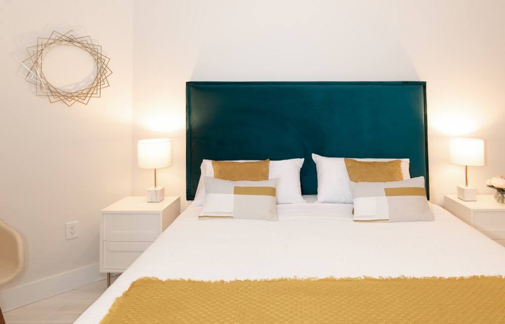 A close-up of the bed in bedroom-1, featuring a teal upholstered headboard, white bedding with yellow throw and decorative pillows, and white nightstands with modern lamps.