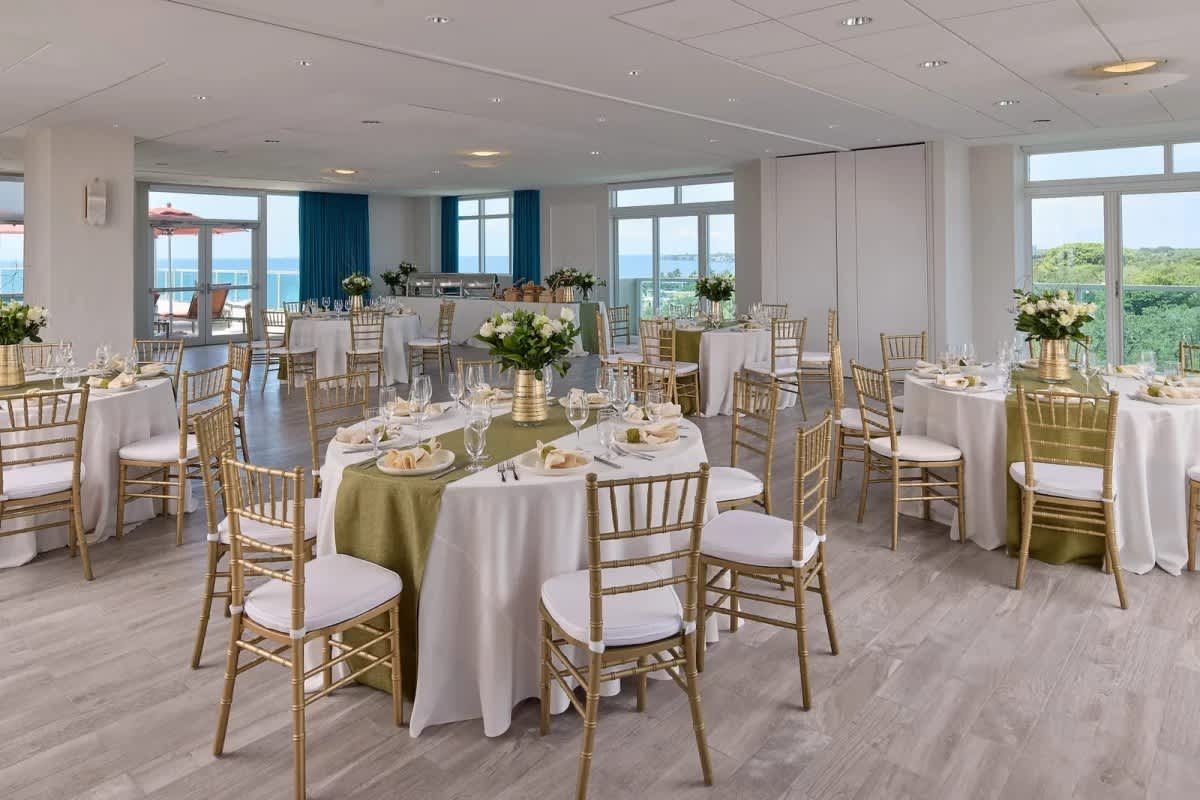 A bright and spacious event space featuring multiple round tables elegantly set with white tablecloths, green runners, and gold chairs, with large windows offering scenic ocean views.