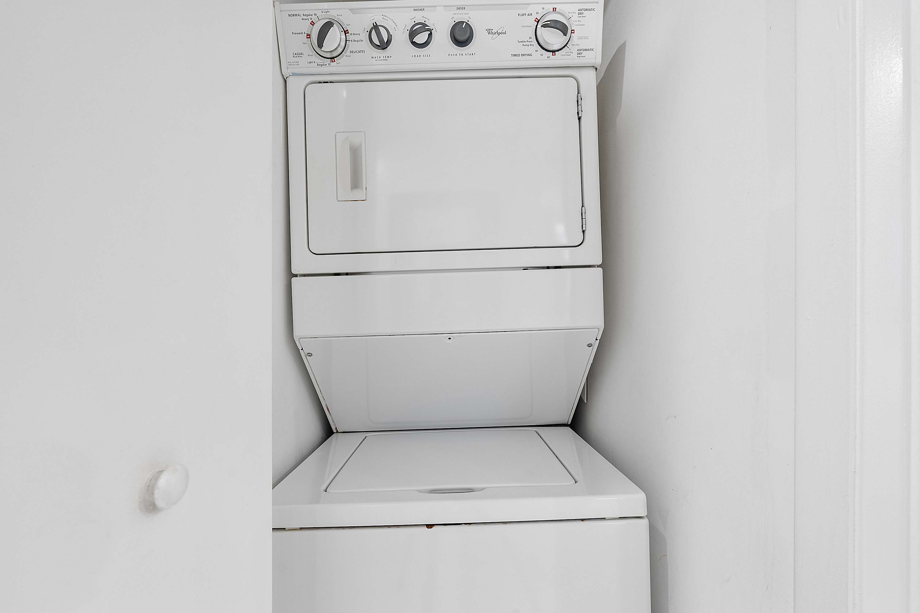 A compact laundry closet houses a stacked white washer and dryer unit, with a simple white door and wall. The appliances appear to be top-loading for the washer and front-loading for the dryer.