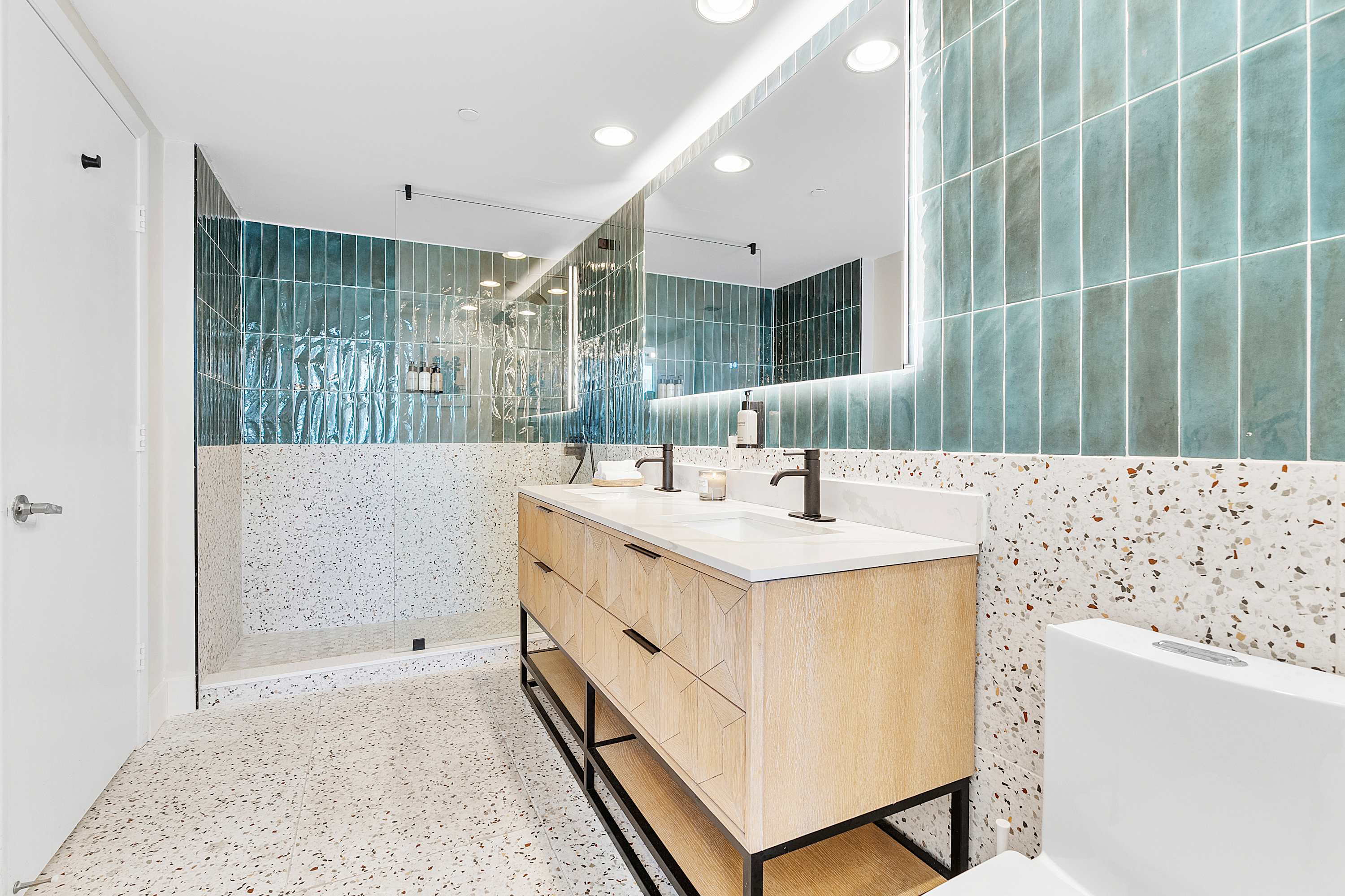 A contemporary bathroom features a double vanity with a light wood cabinet and black hardware, set against a white terrazzo-style floor and wall. A walk-in shower with teal subway tiles and a glass enclosure is visible.