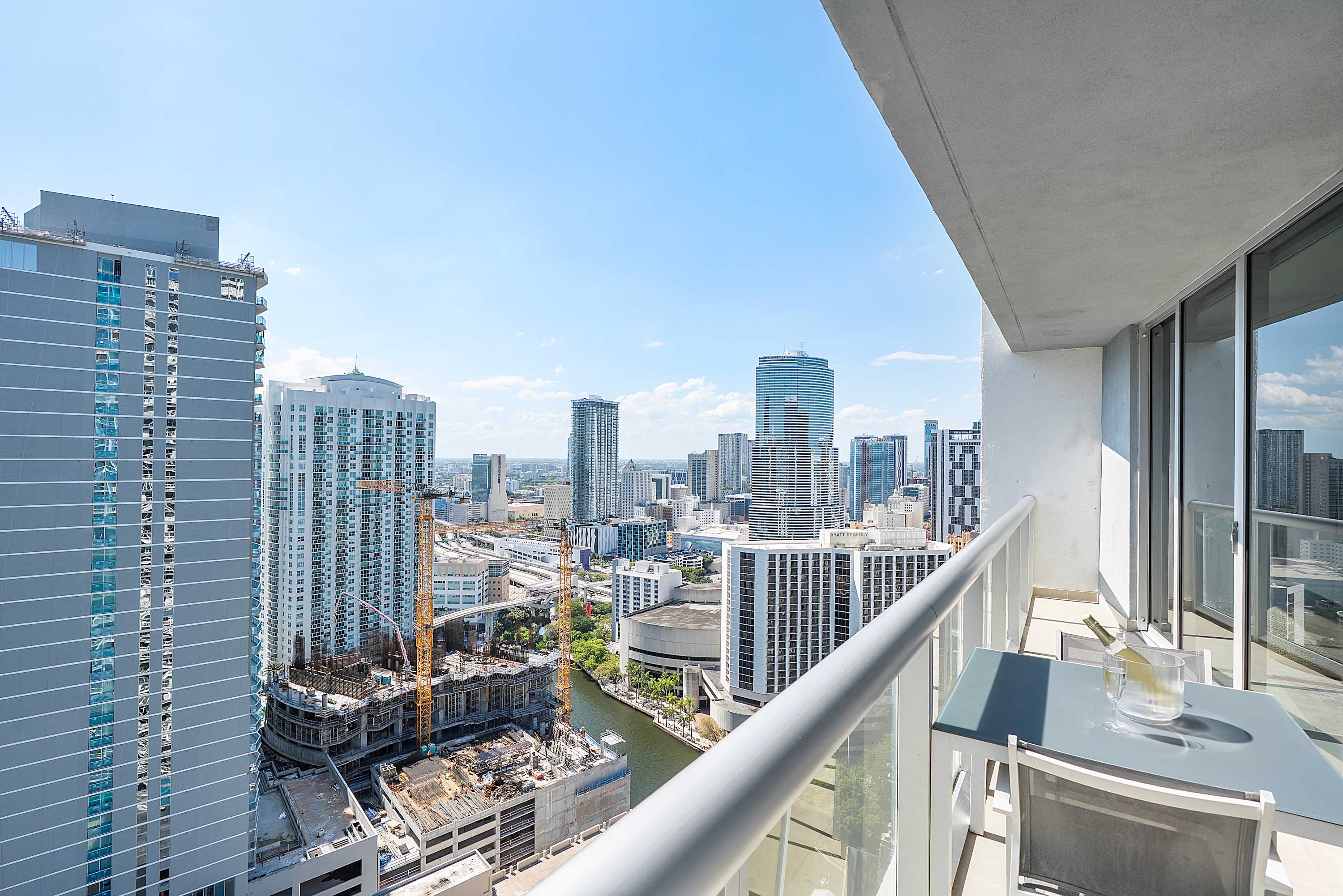 High-rise balcony with a small outdoor table and two chairs, offering expansive city and waterfront views with numerous modern buildings and construction sites under a clear blue sky.