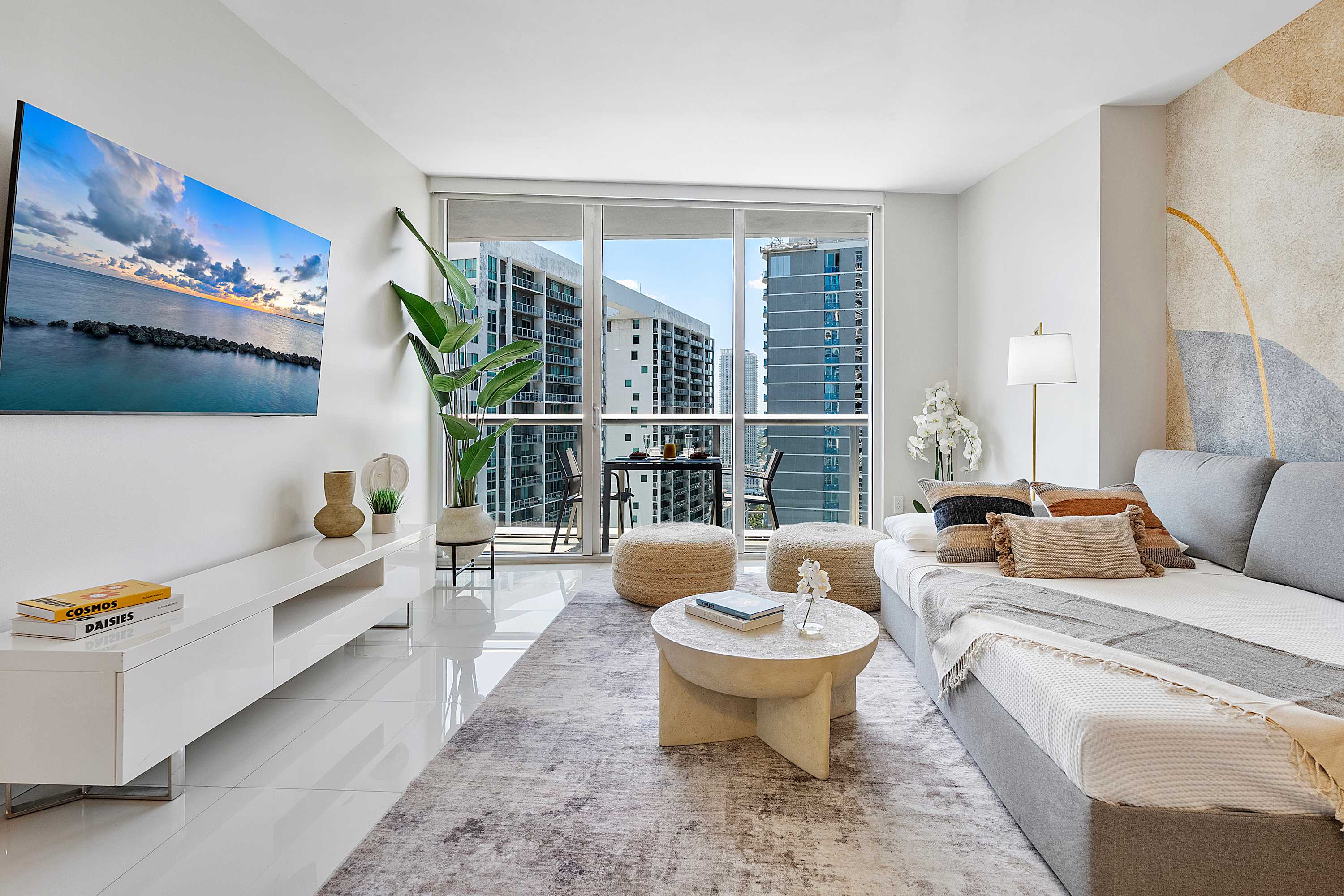 Modern living room with a grey sectional sofa, a round coffee table, and a wall-mounted TV, featuring floor-to-ceiling windows with city views and a decorative accent wall with abstract patterns.