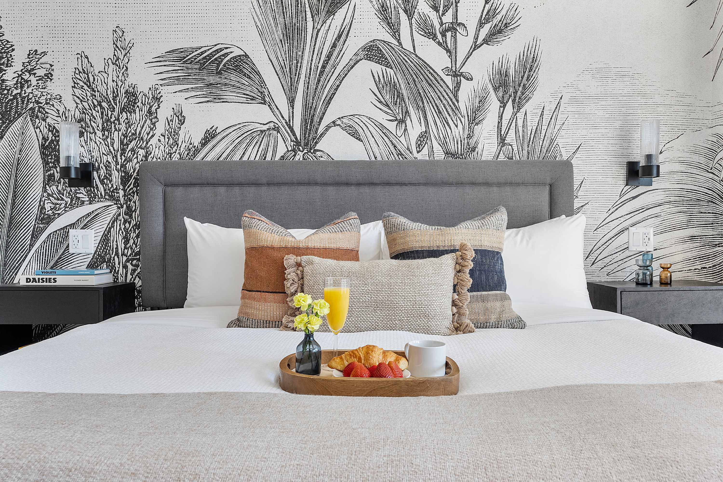 Cozy bedroom featuring a large bed with a grey upholstered headboard, decorative pillows, and a breakfast tray with fruit and juice, set against a striking black and white botanical wallpaper.